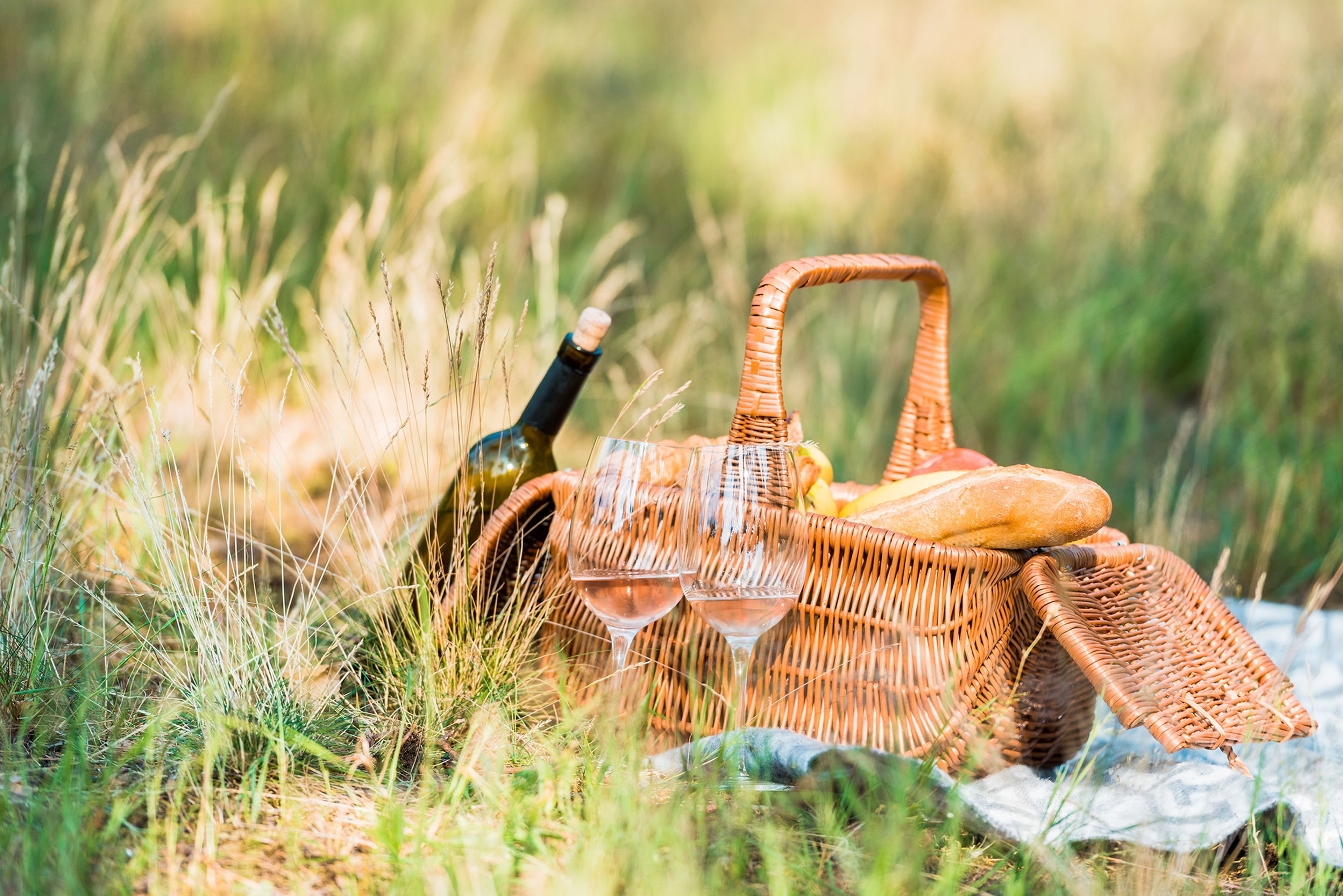 Ein geflochtener Picknickkorb mit Wein, zwei Gläsern, Brot und Obst auf einer Decke auf einer sonnigen Wiese.