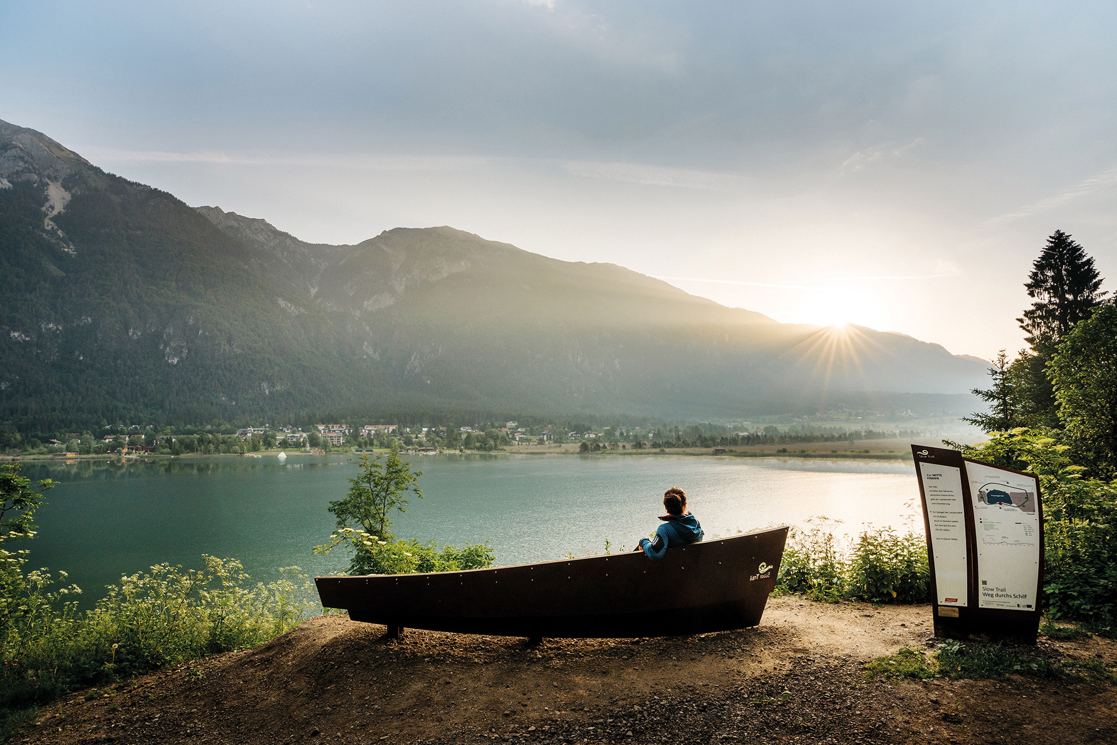 Person, die auf einer Bank in Form eines Bootes sitzt und bei Sonnenaufgang auf den Pressgger See und die Berge blickt. In der Nähe befindet sich ein Informationsschild.
