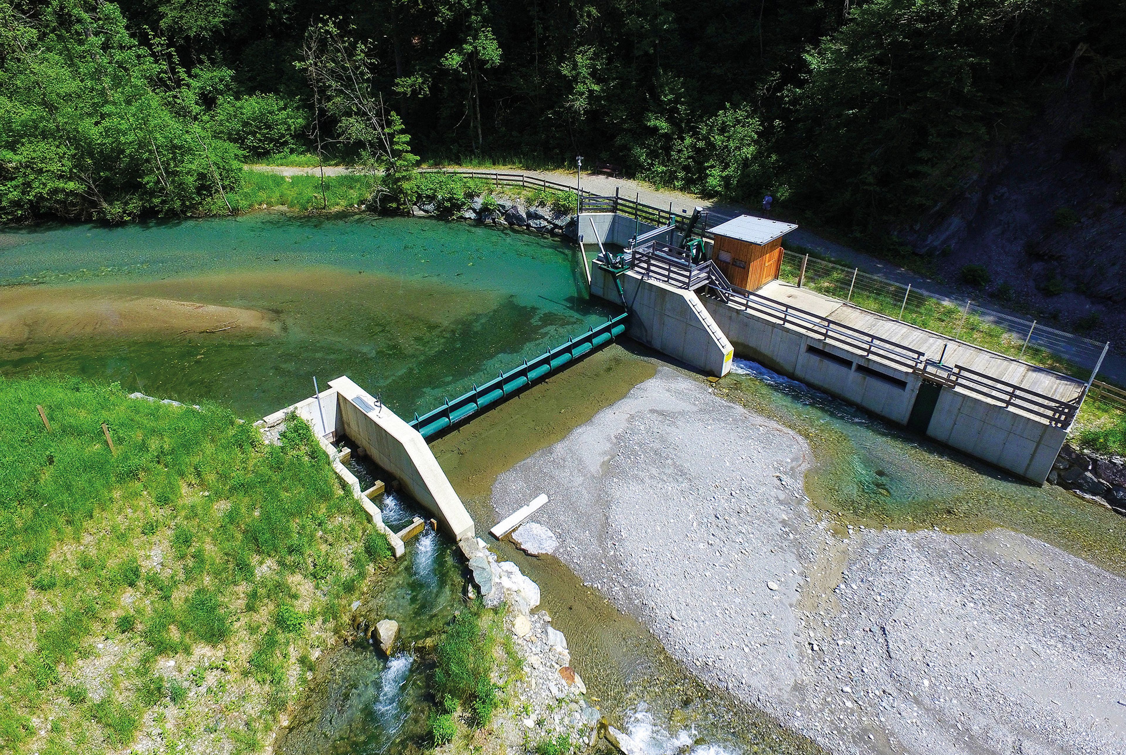 Luftaufnahme auf das Wasserkraftwerk des Schloss Lerchenhof mit einem kleinen Staudamm mit klarem blaugrünem Wasser, umgeben von üppigem Grün und einer Kiesbereich, mit einem Holzschuppen in der Nähe.