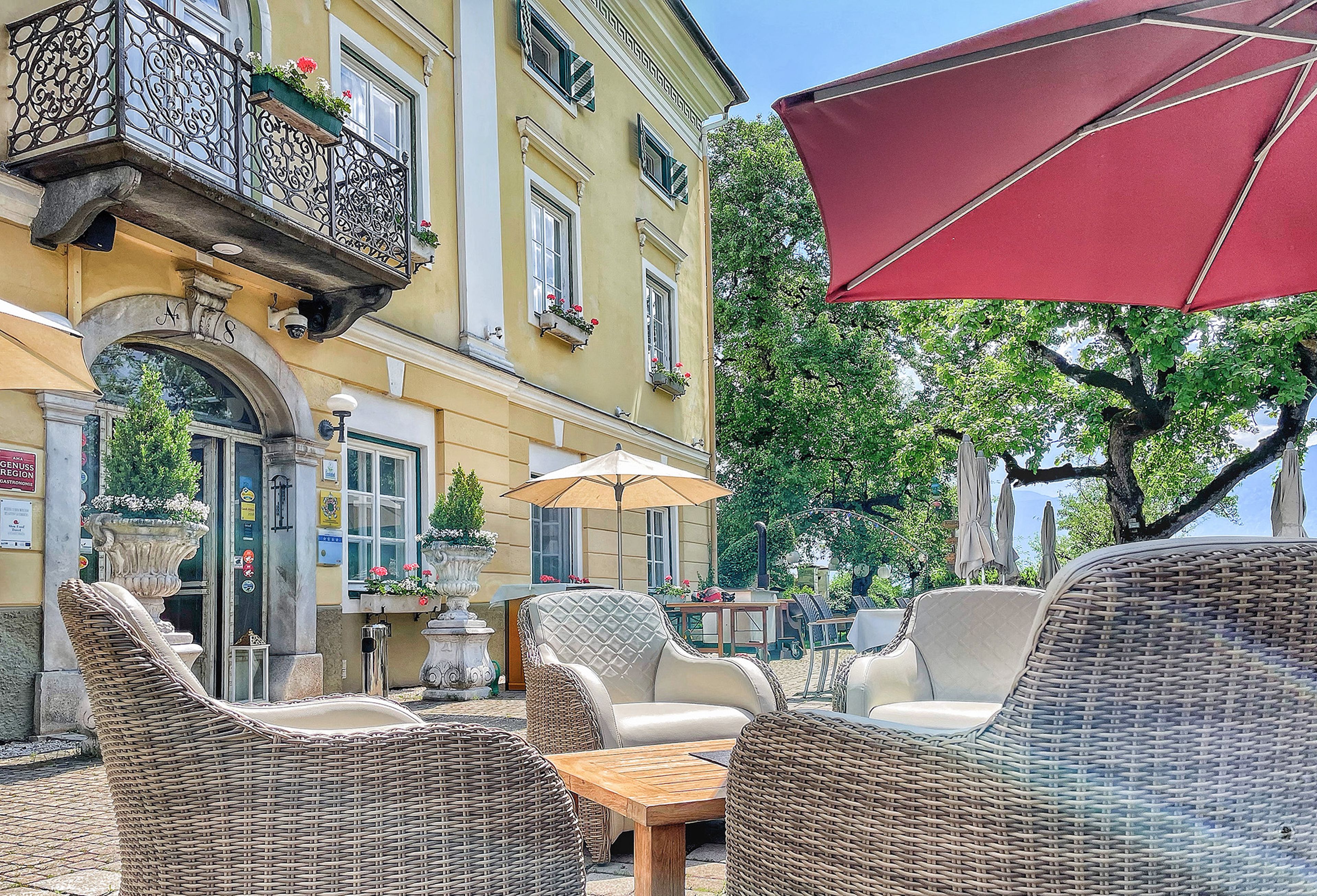 Die Außenterrasse des Hotel und Restaurants Schloss Lerchenhof mit bequemen Korbstühlen und Sonnenschirmen umgeben von Bäumen.