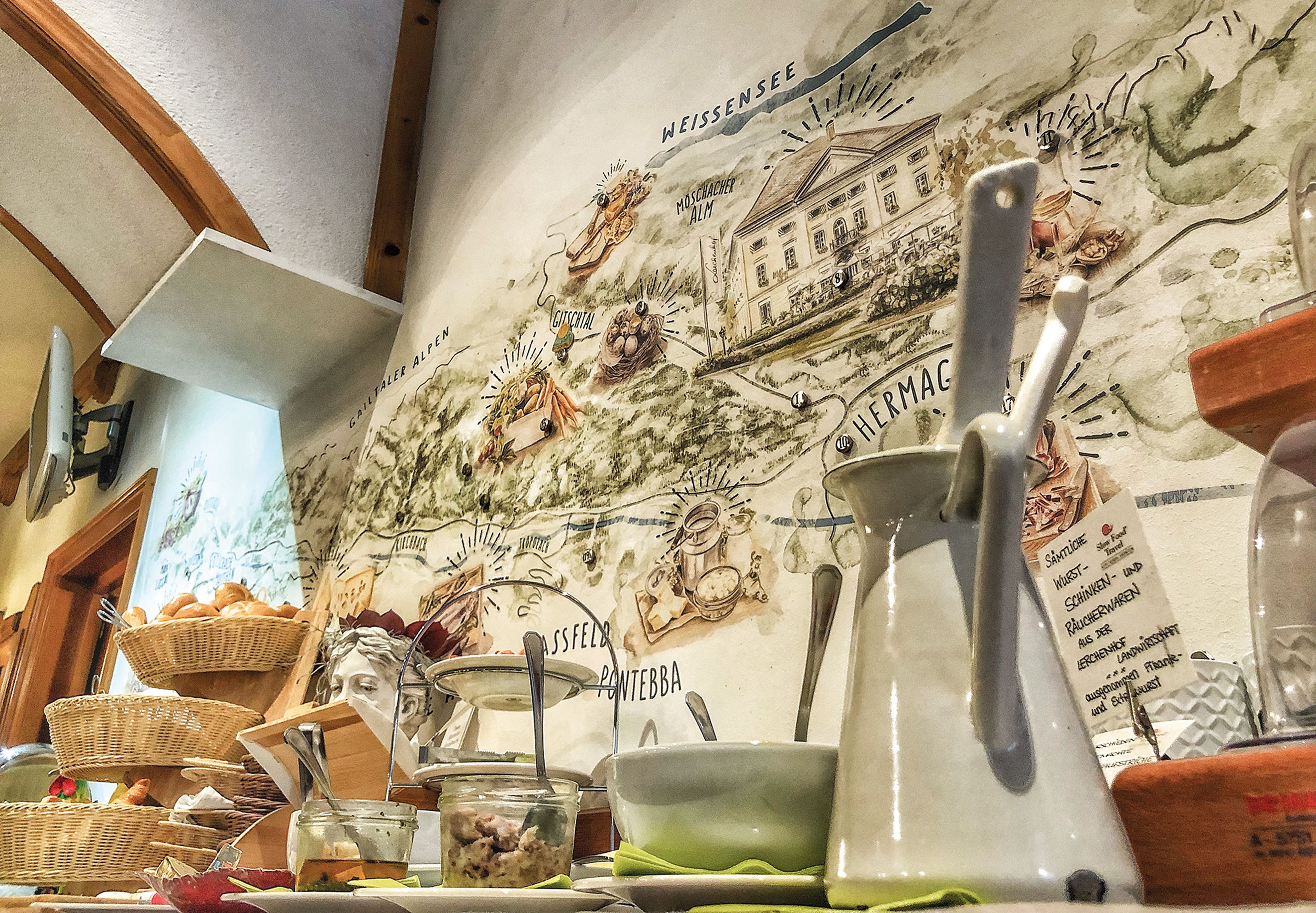 Ein Frühstücksbuffet mit Brotkörben, Gläsern und Besteck vor einer dekorativen Wandkarte mit Illustrationen und Text.