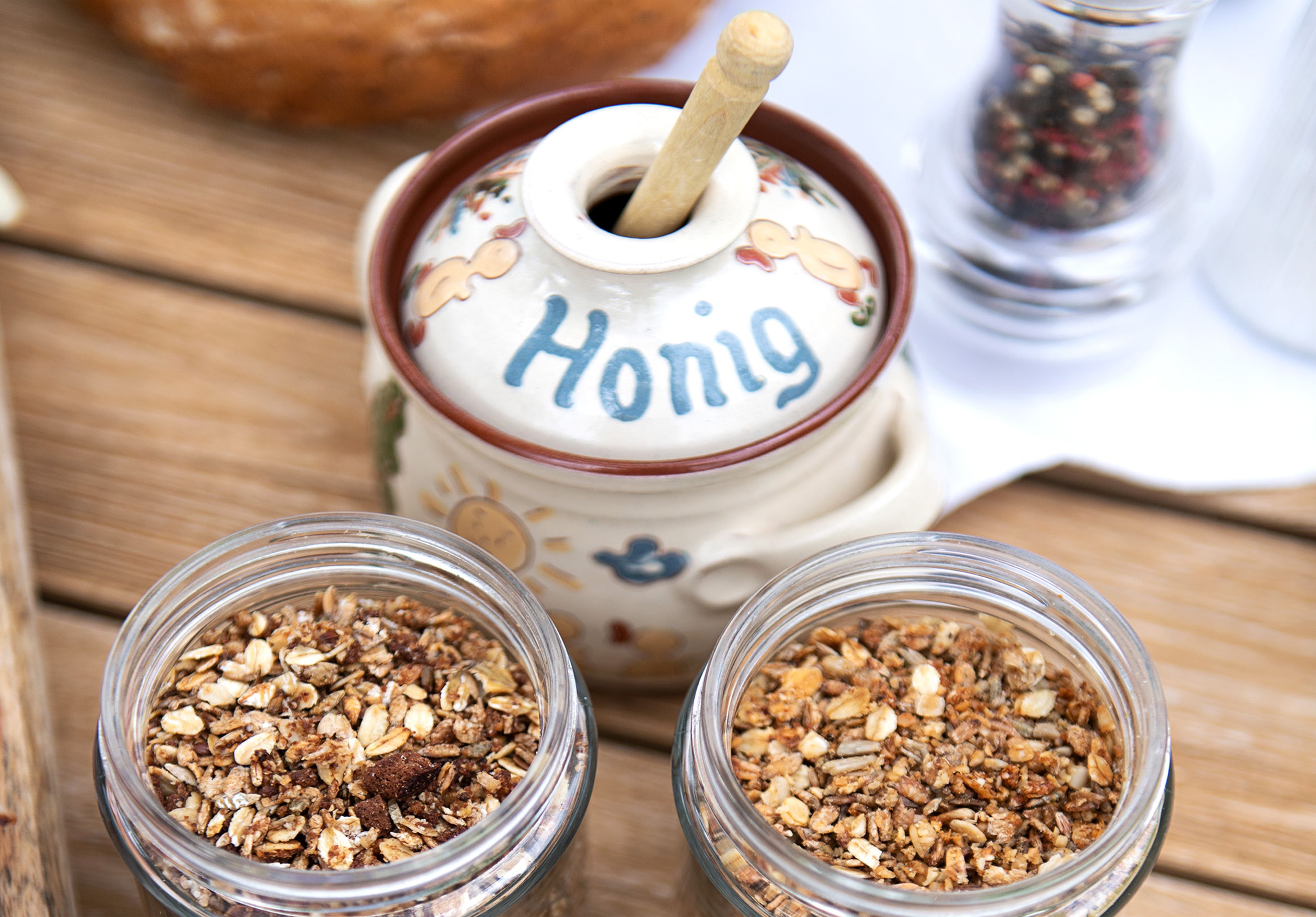 Honigtopf aus Keramik mit der Aufschrift „Honig“, umgeben von Müsligläsern auf einem Holztisch.