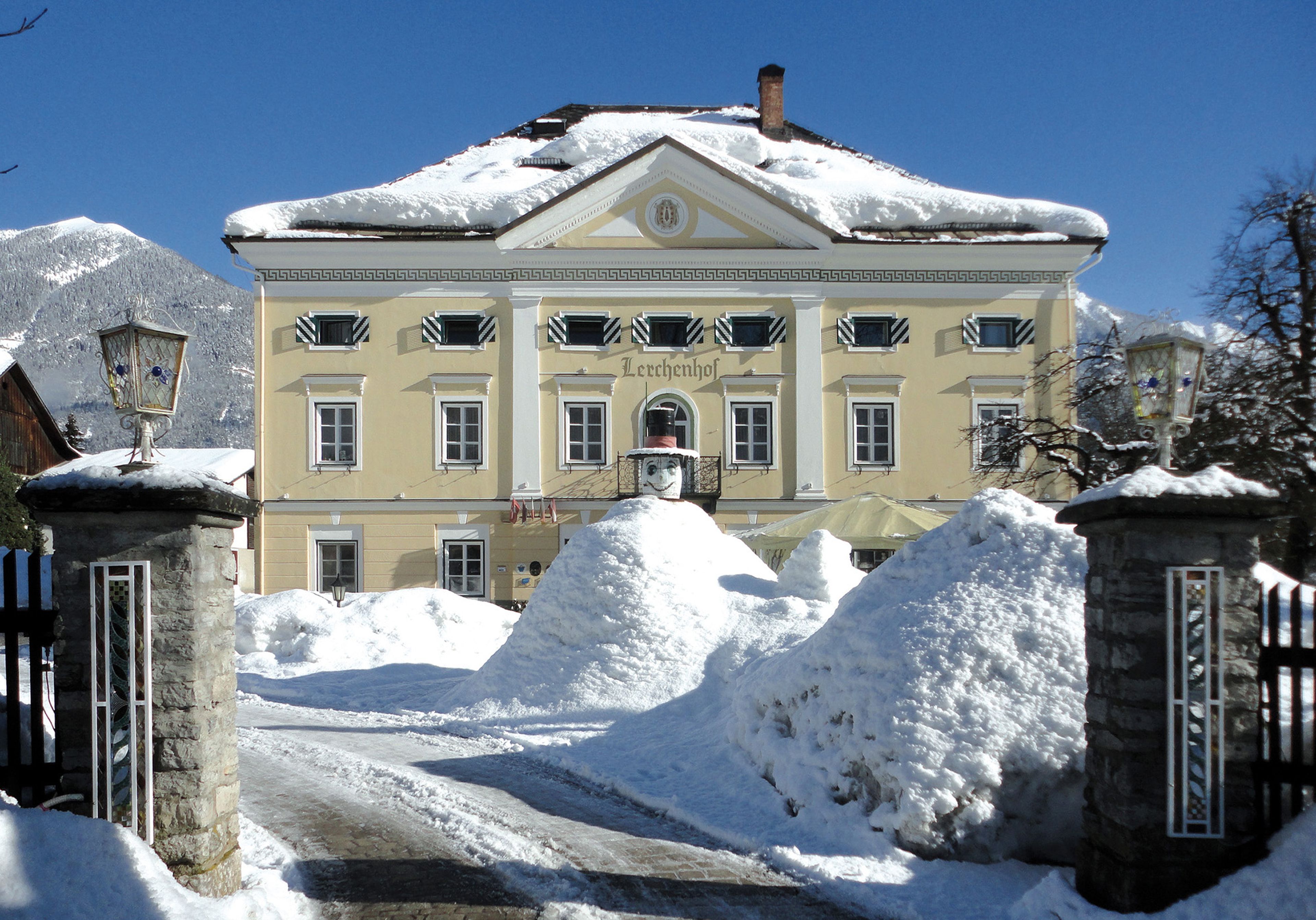 Das große, schneebedeckte Schloss Lerchenhof mit gelber Fassade vor der Kulisse schneebedeckter Berge und eines klaren blauen Himmels.