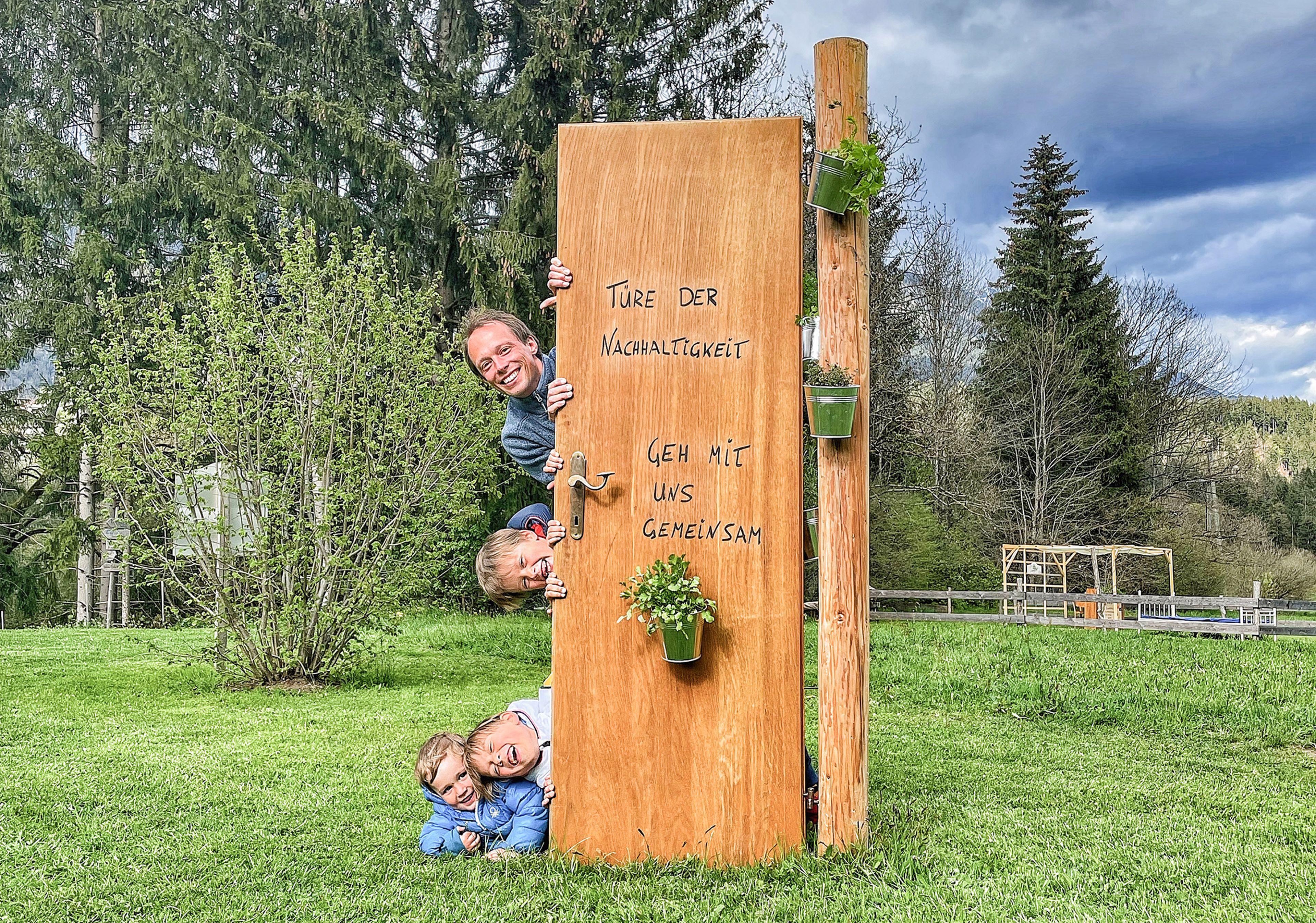 Johann Steinwender mit seinen Söhnen spähen durch eine Holztür mit der Aufschrift „Türe der Nachhaltigkeit“ in eine Rasenfläche mit Bäumen und bewölktem Himmel.