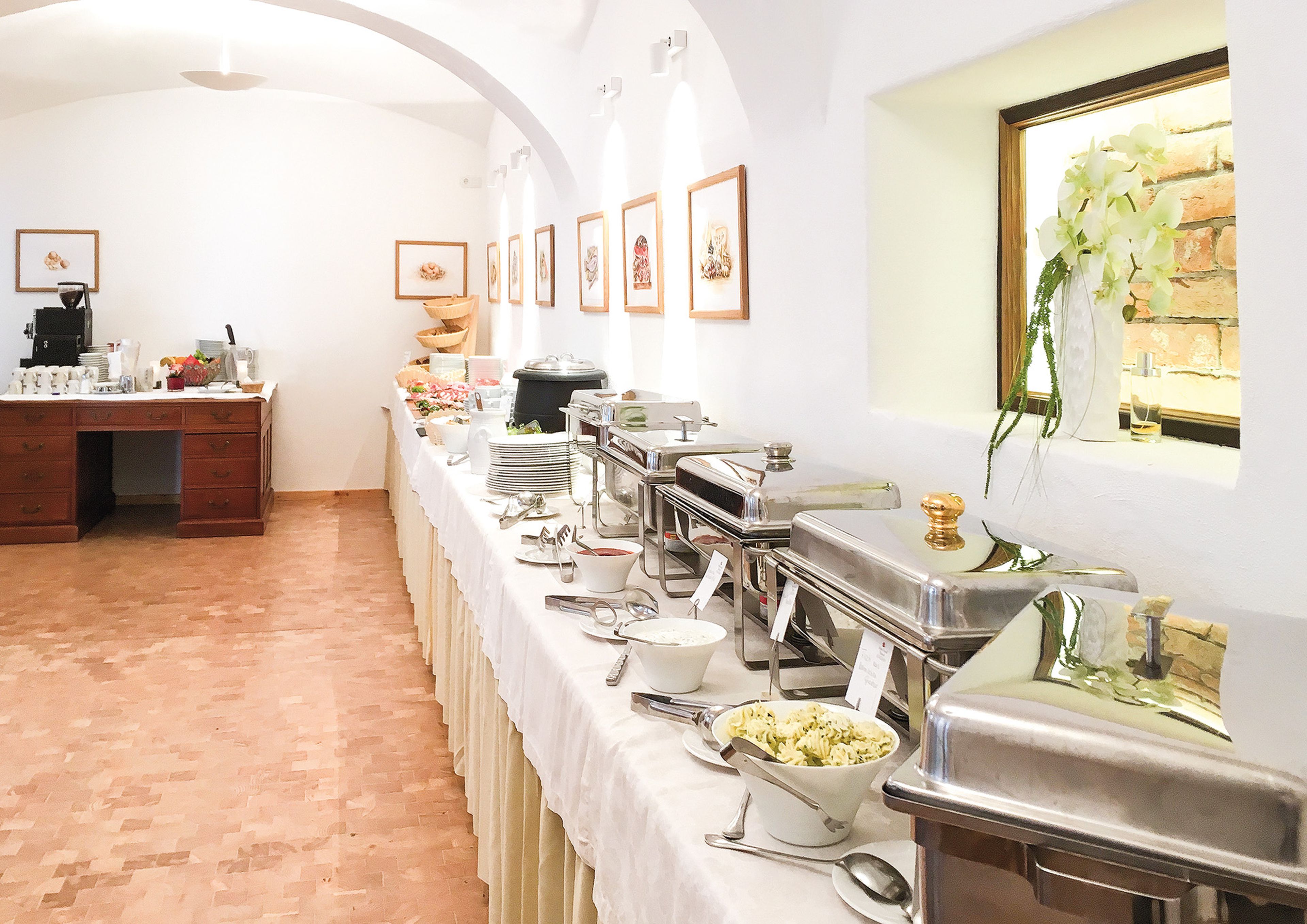 A buffet setup in a bright room with chafing dishes, plates, and salads on a long table, and framed art on the wall.