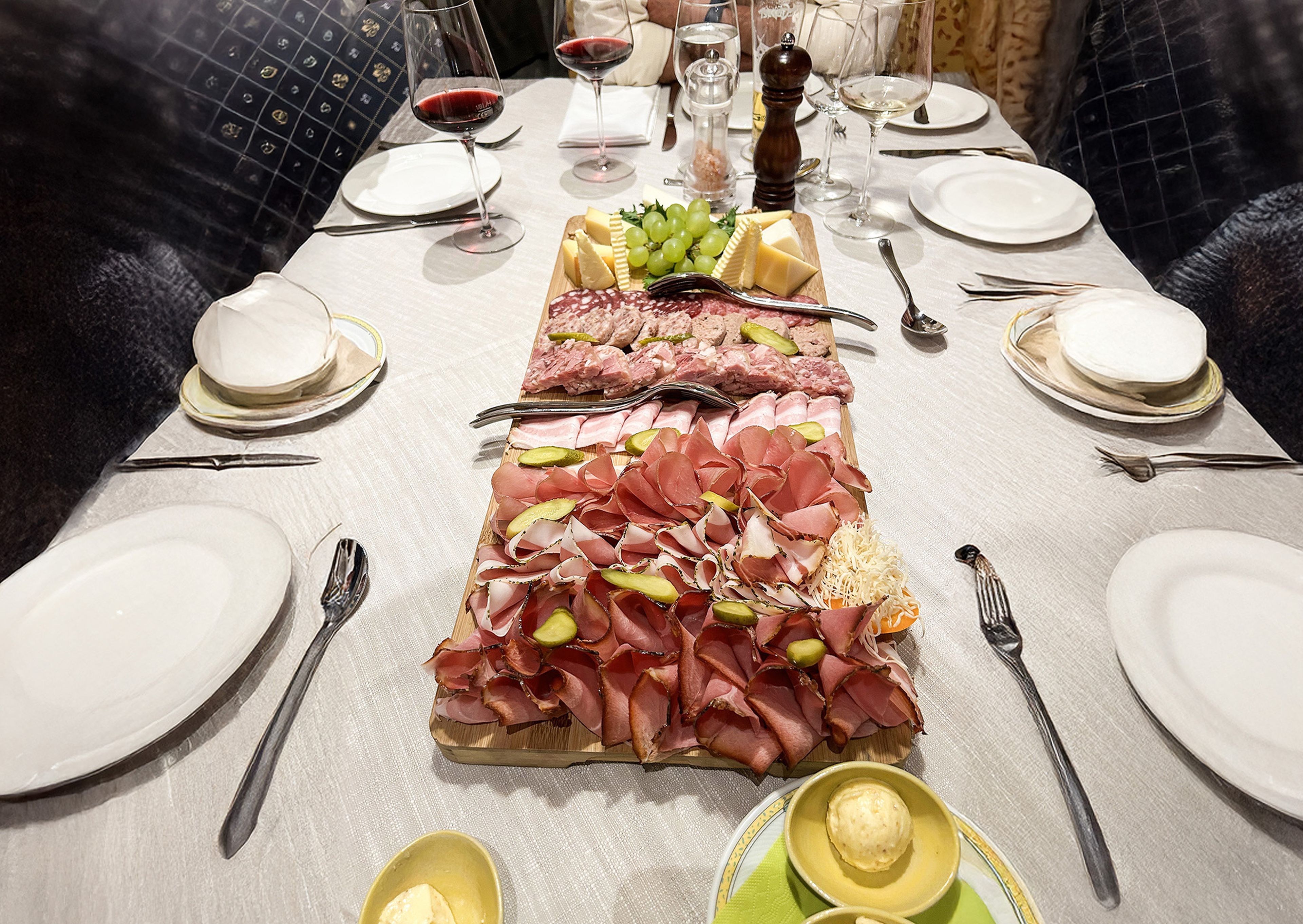 A dining table set for six with plates, cutlery, red wine, and a large platter of assorted meats, cheeses, and grapes in the center.