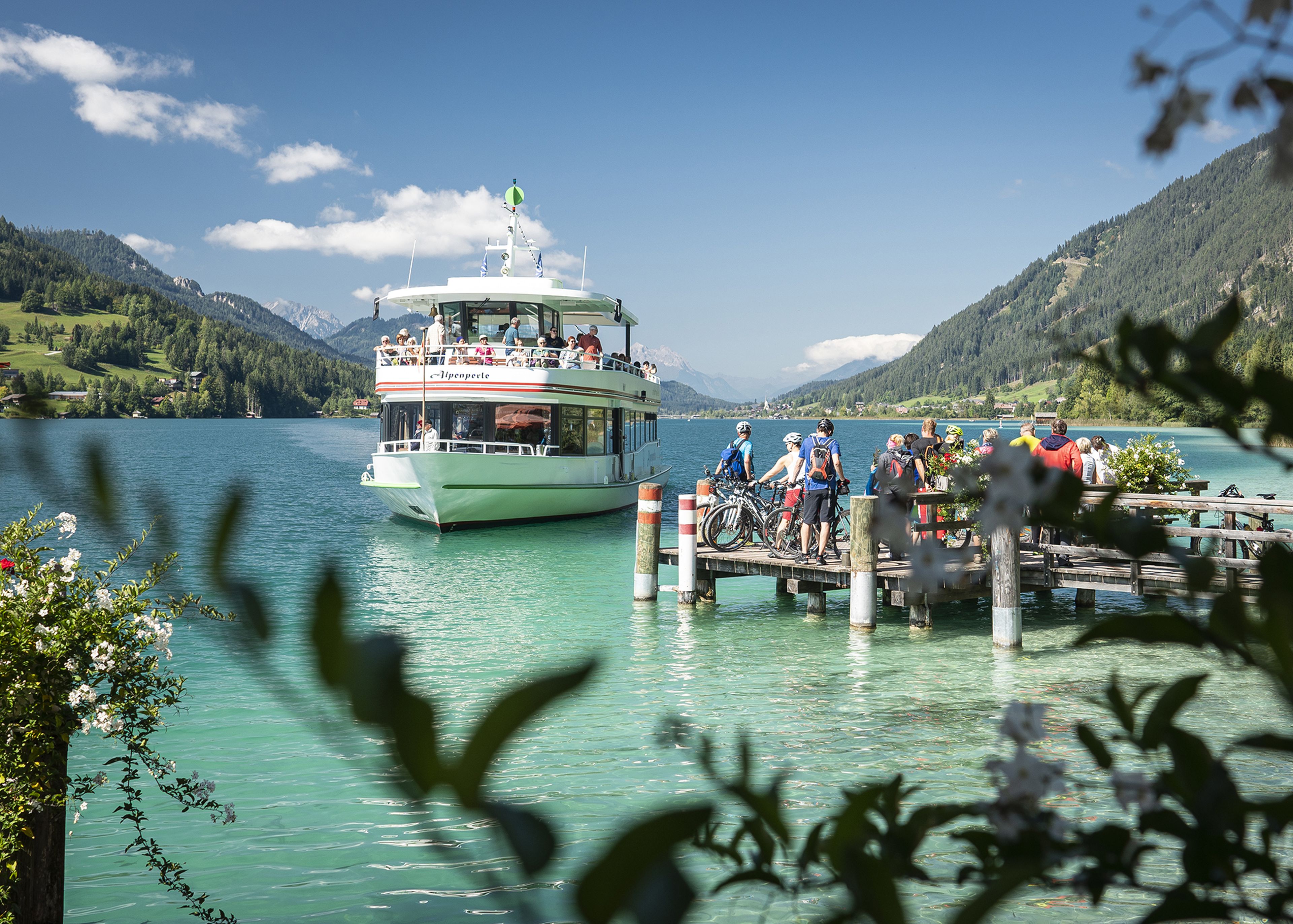 Ein Boot nähert sich einer Anlegestelle an einem türkisfarbenen See, der von Bergen umgeben ist. Menschen mit Fahrrädern warten am Steg unter einem klaren blauen Himmel.