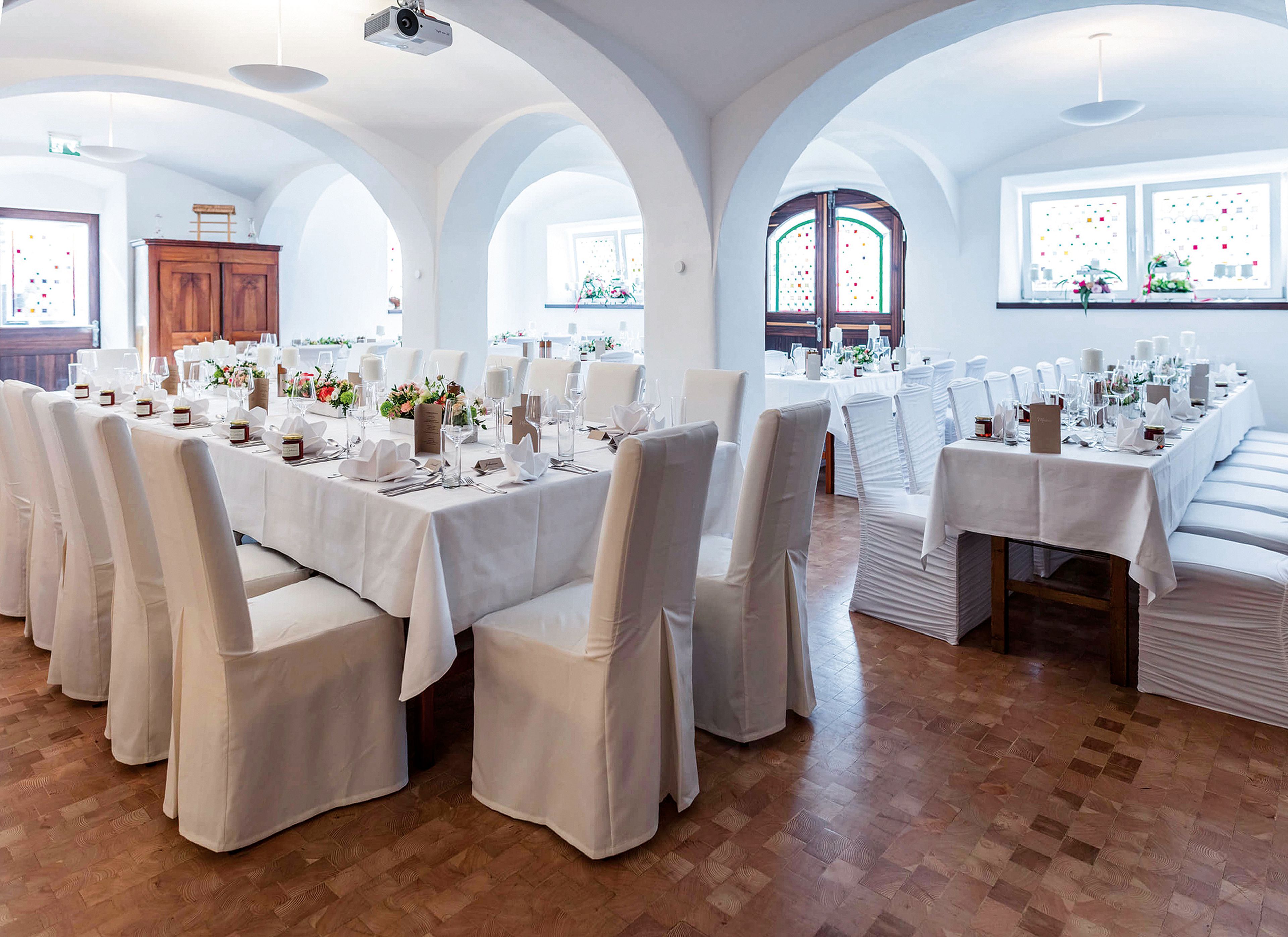 Eleganter, festlich gedeckter Bankettsaal mit weißen Tischdecken und Stuhlhussen, dekoriert mit Blumenarrangements und Kerzen, in einem hellen Raum mit gewölbten Decken und bunten Fenstern.
