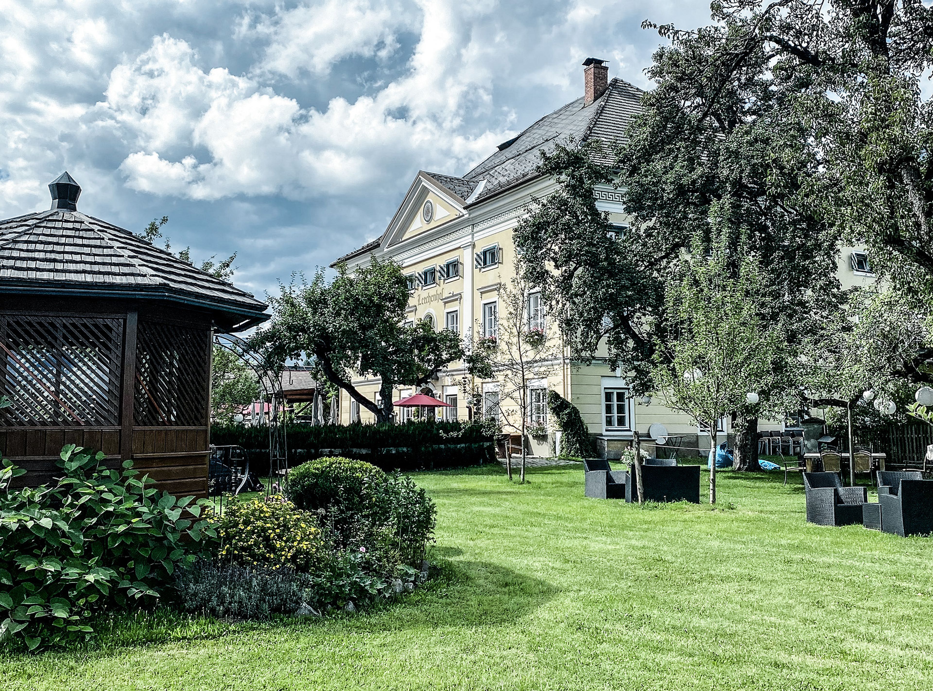 Das Schloss Lerchenhof – ein großes, elegantes Gebäude mit einem schrägen Dach steht neben einem üppigen Garten mit einem Pavillon, Bäumen und Sitzgelegenheiten im Freien unter einem bewölkten Himmel.