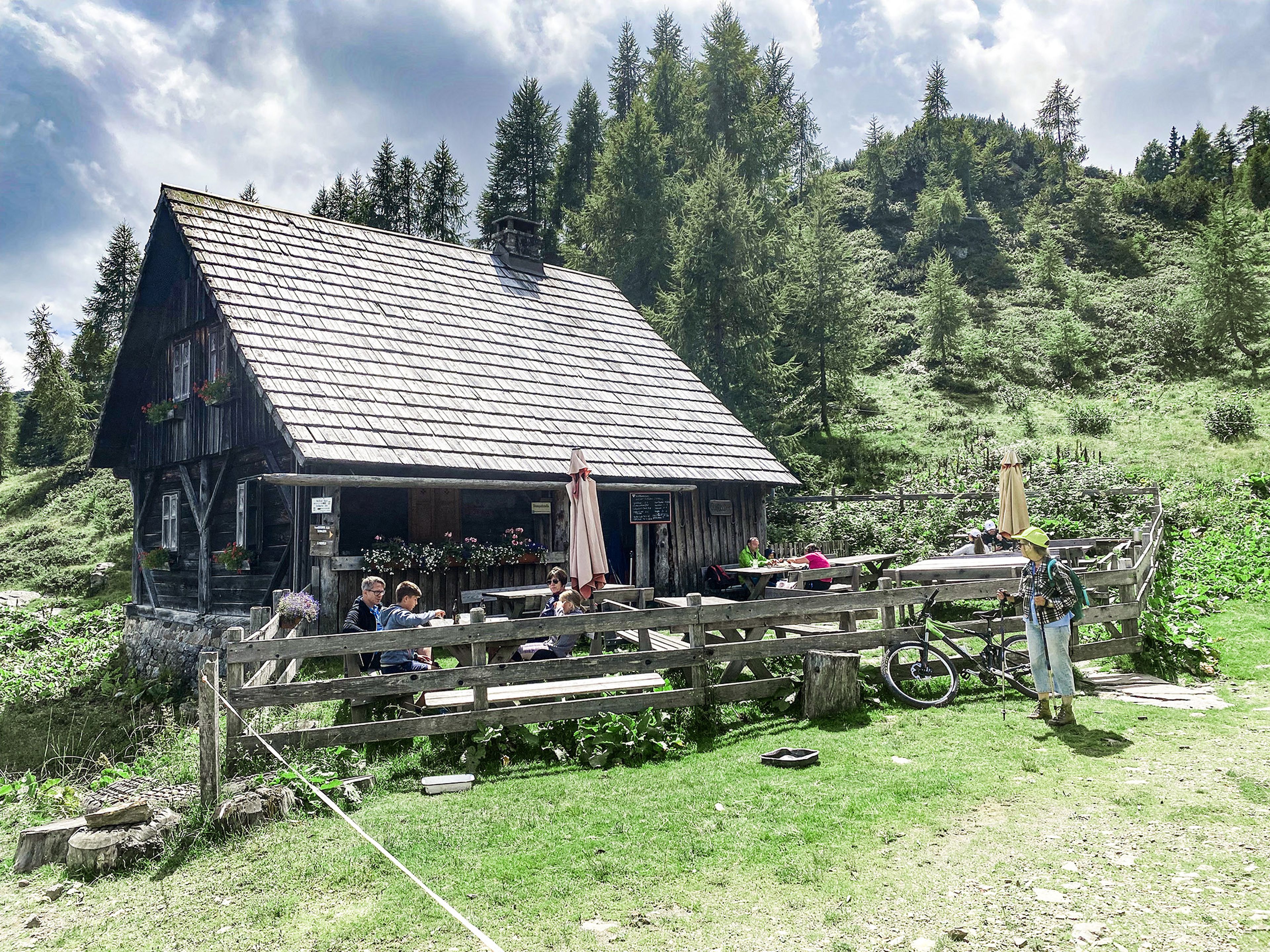 Eine rustikale Hütte mit Holzzaun in einer üppigen Hügellandschaft. Menschen sitzen an Tischen im Freien, ein Fahrrad lehnt am Zaun.