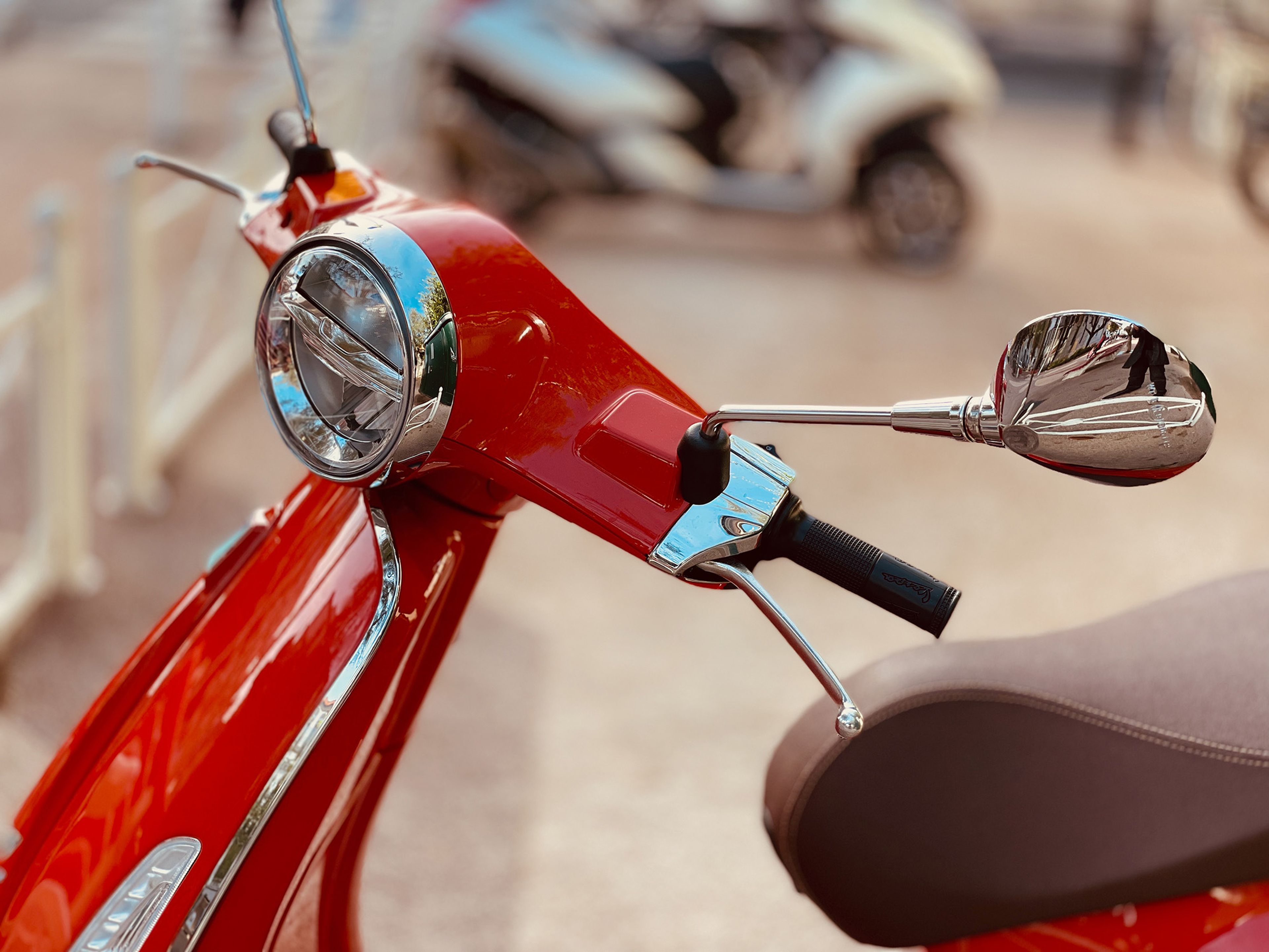 Nahaufnahme des Lenkers und des Spiegels eines roten Motorrollers, mit einem unscharfen Hintergrund eines anderen Motorrollers und einem weißen Zaun.