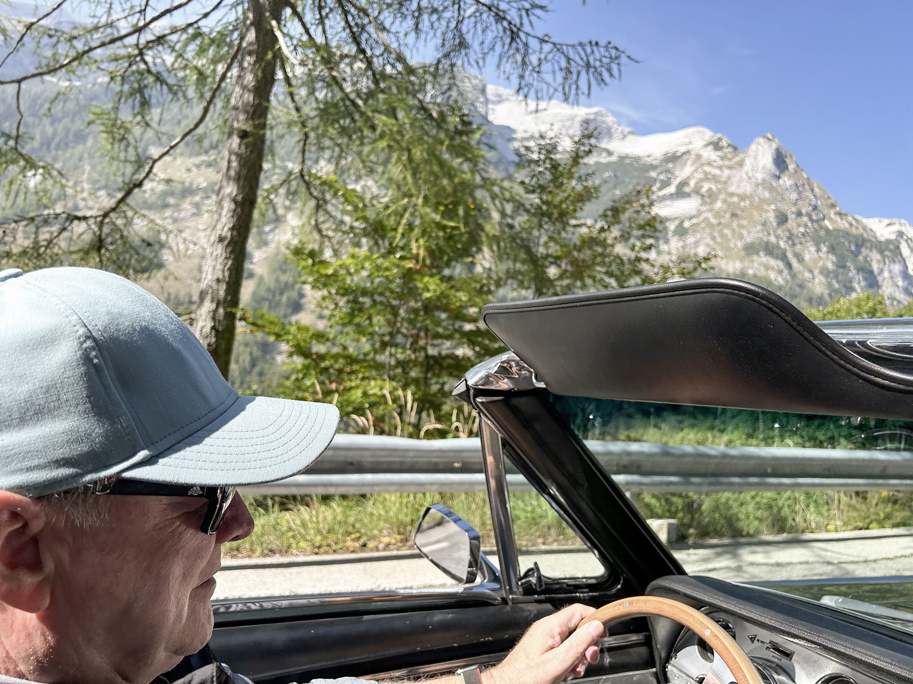 Mann fährt ein Cabrio auf einer malerischen Bergstraße, trägt eine Mütze und eine Sonnenbrille, im Hintergrund sind Bäume und schneebedeckte Gipfel zu sehen.