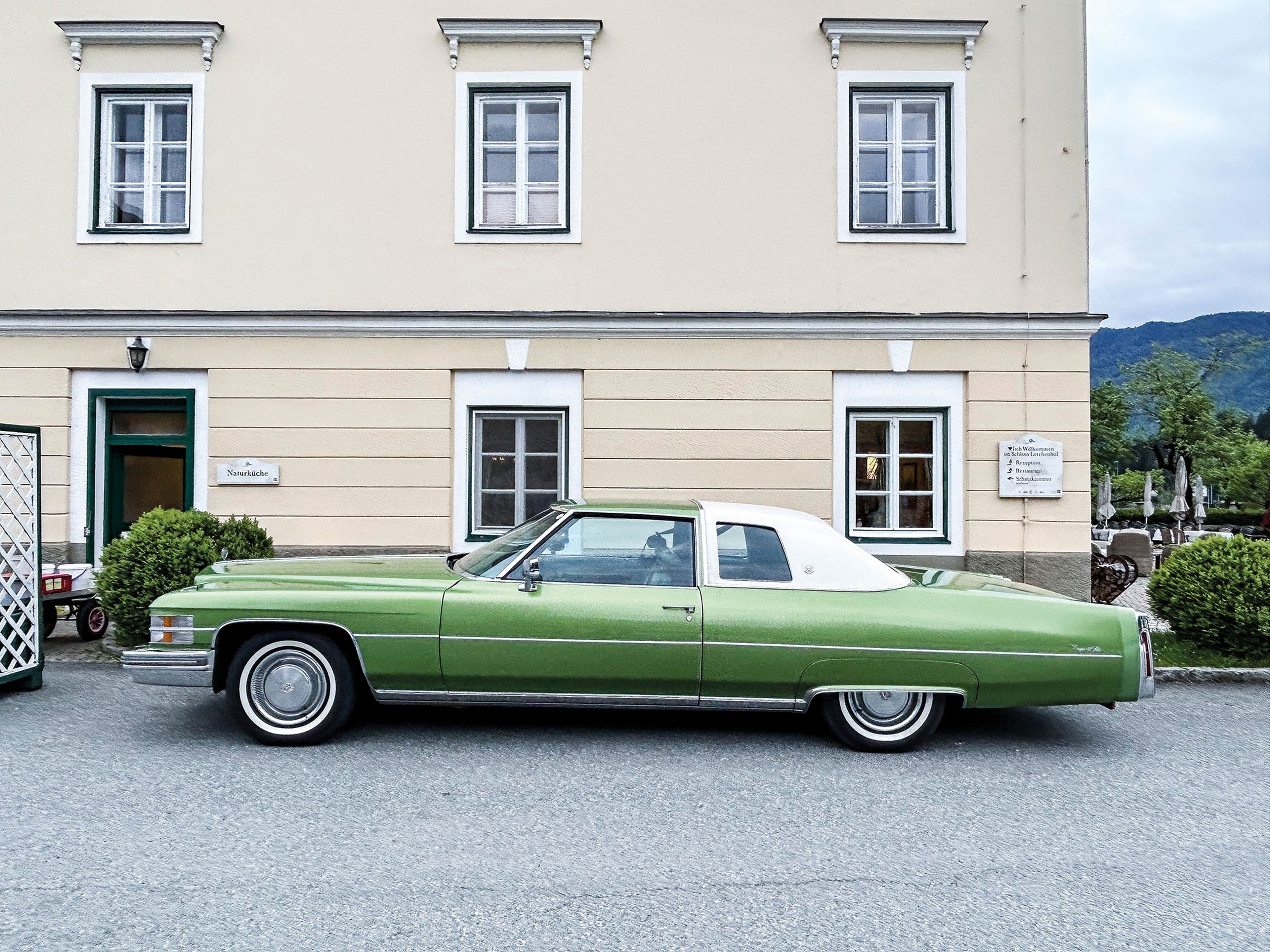 A vintage green car with a white roof is parked in front of a beige building with four windows, set against a mountainous backdrop.
