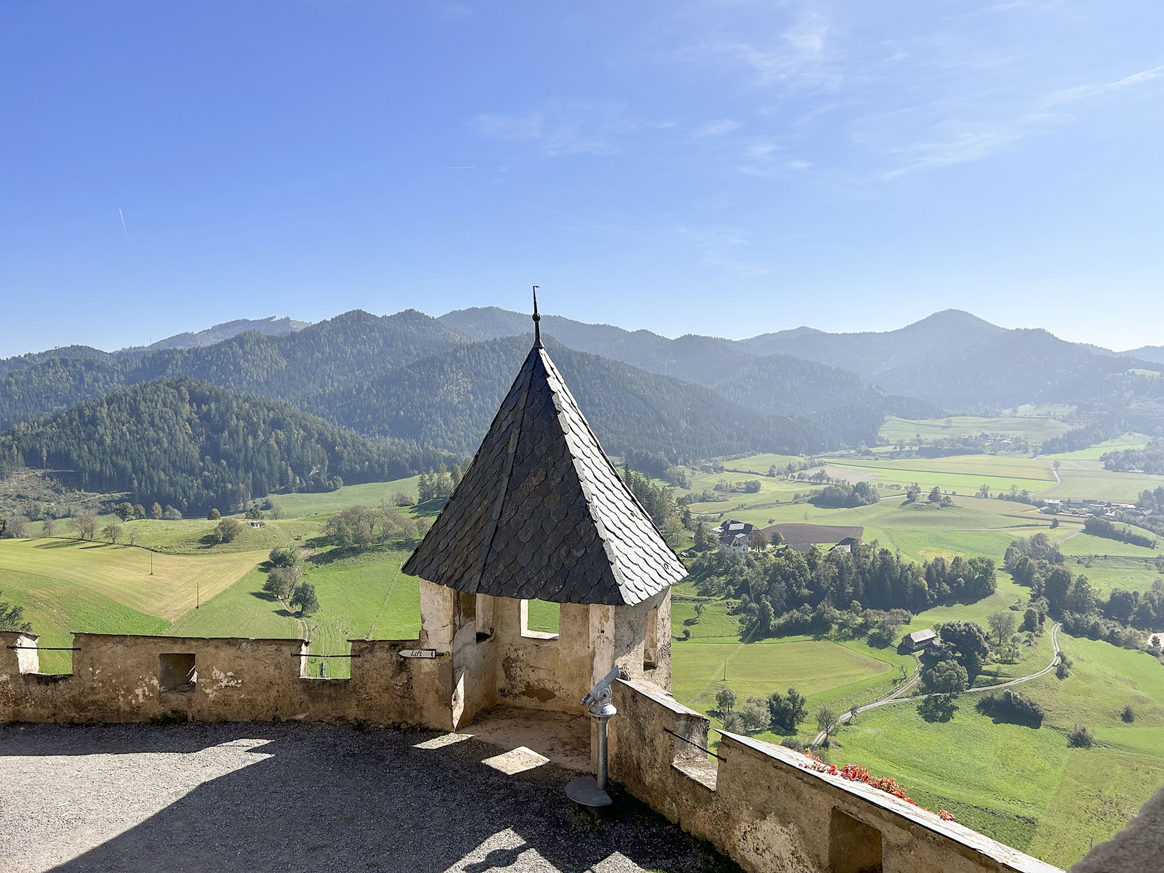Ein kleiner Steinturm überblickt ein weites, grünes Tal mit sanften Hügeln und vereinzelten Bäumen unter einem klaren blauen Himmel.