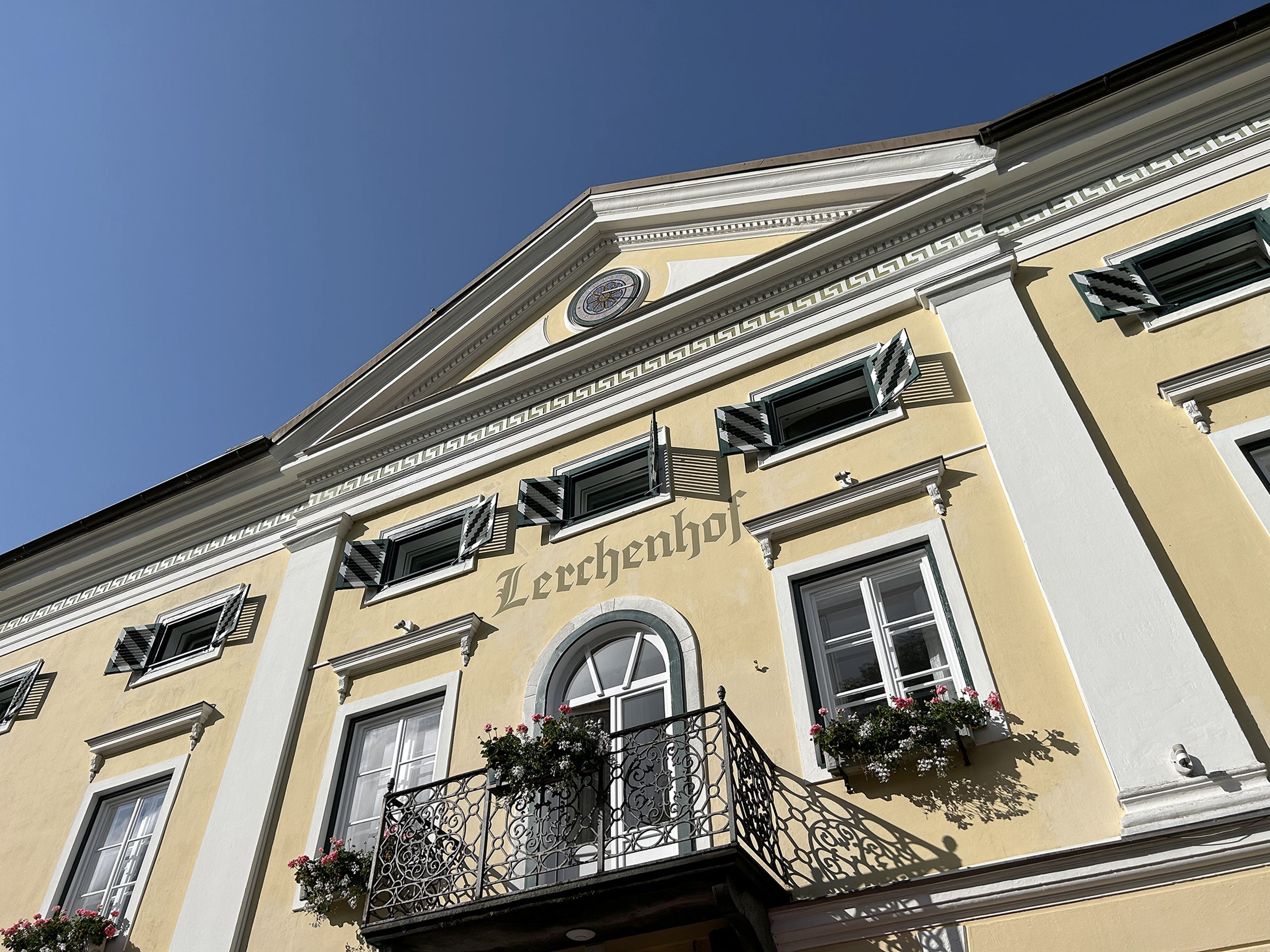 Gelbes Gebäude mit grünen Fensterläden, einem Balkon mit Blumenkästen und der Aufschrift „Lerchenhof“ über dem Bogenfenster unter einem strahlend blauen Himmel.