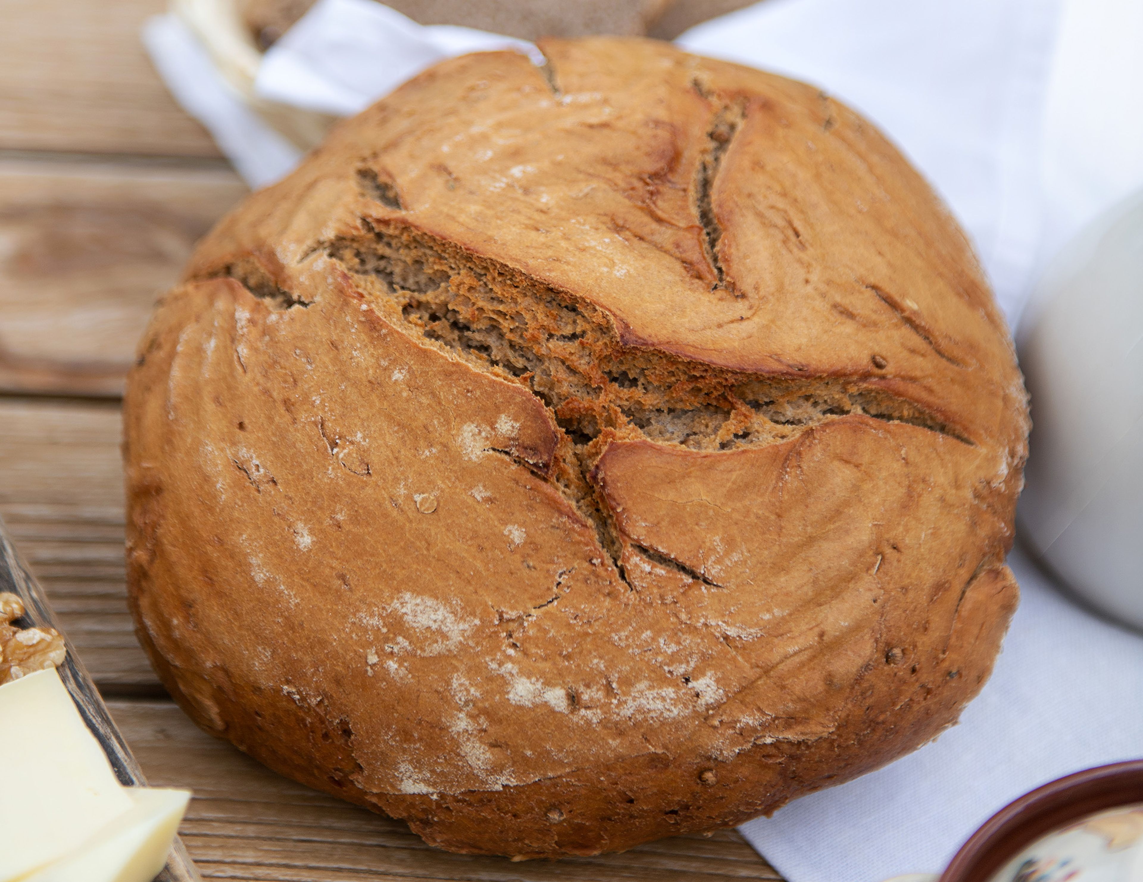 Ein runder, knuspriger Laib Brot mit rissiger Oberfläche liegt auf einer Holzoberfläche, umgeben von einem weißen Tuch und einem Blick auf ein Schneidebrett.