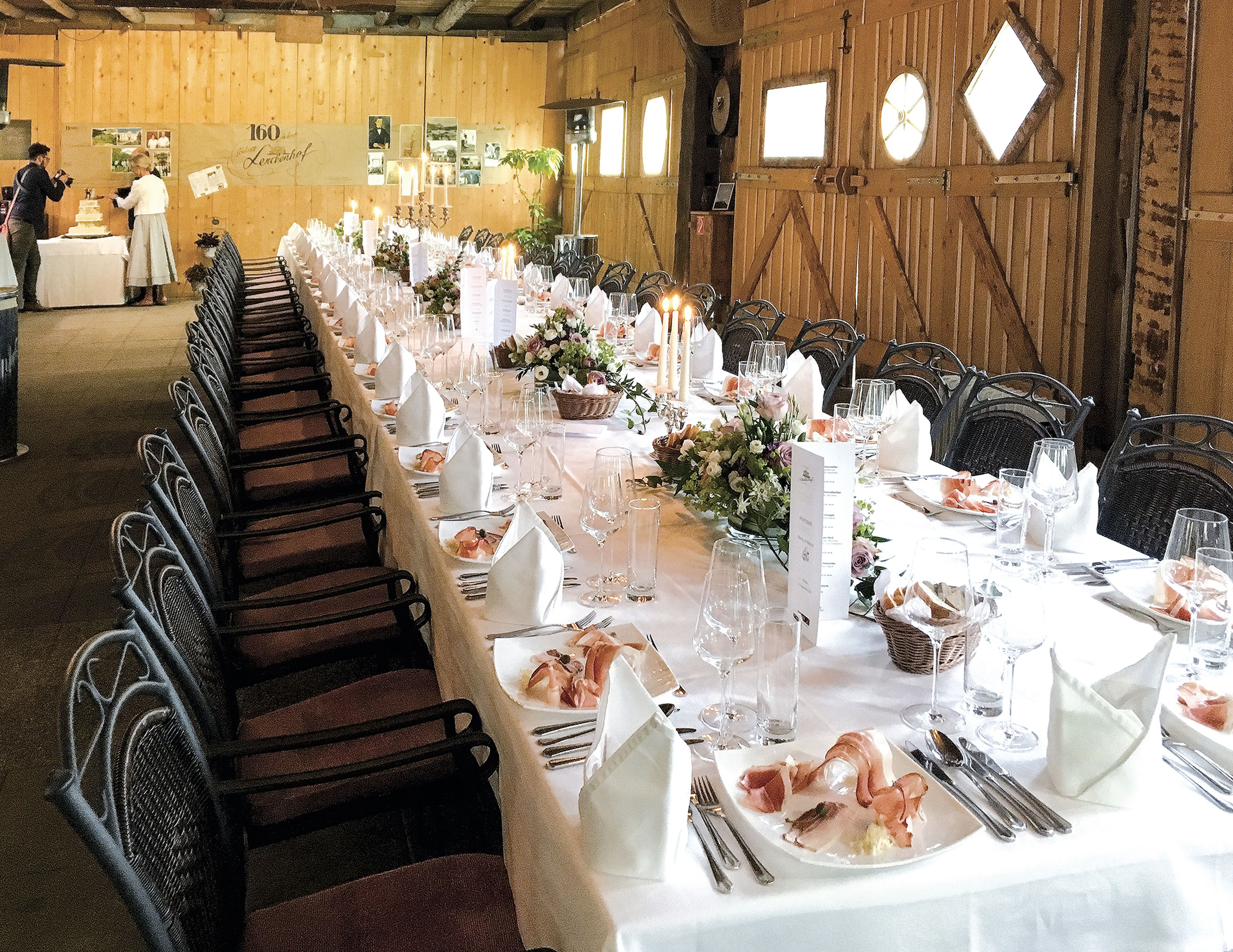 Langer Esstisch, gedeckt für ein Bankett in einem rustikalen Holzsaal, geschmückt mit Blumengestecken, Kerzen und ordentlich arrangiertem Geschirr.