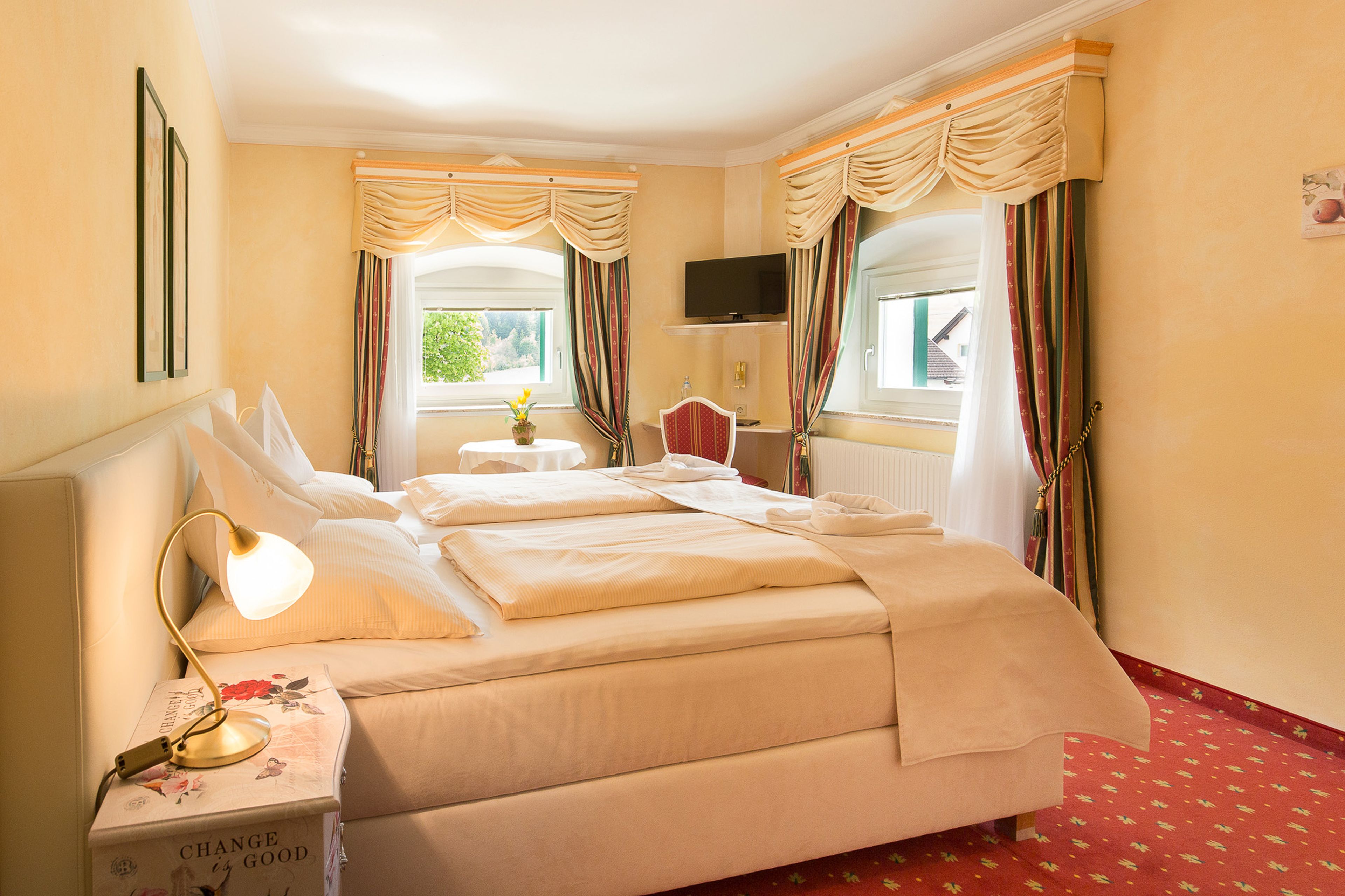 Elegantes Hotelzimmer mit ordentlich gemachtem Doppelbett, Nachttisch mit Lampe, kleiner Essecke und Fenstern mit drapierten Vorhängen.