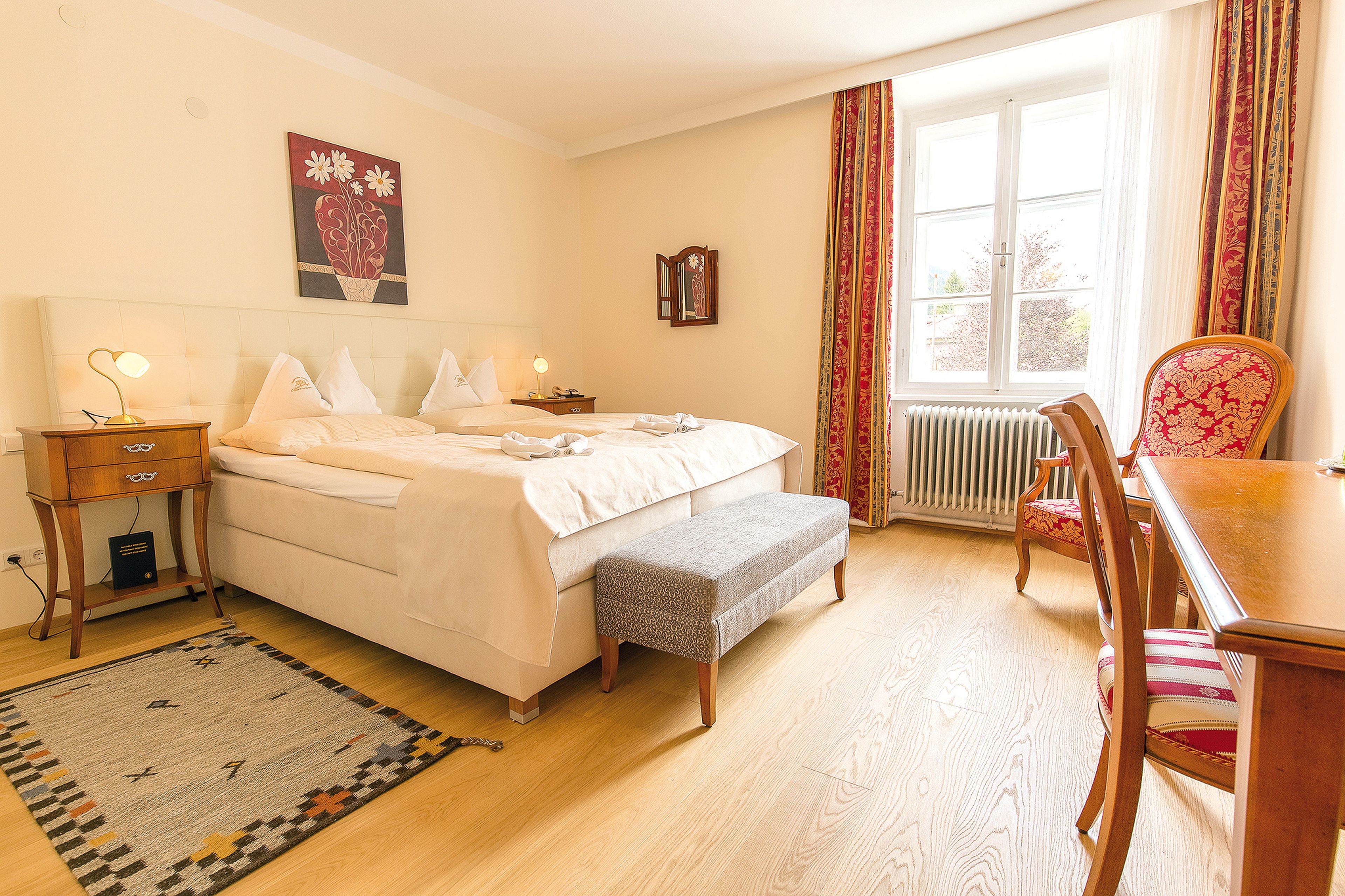 Gemütliches Hotelzimmer mit Doppelbett, Holzmöbeln, einem dekorativen Teppich und einem Fenster mit roten Vorhängen, durch die natürliches Licht den Raum erhellt.