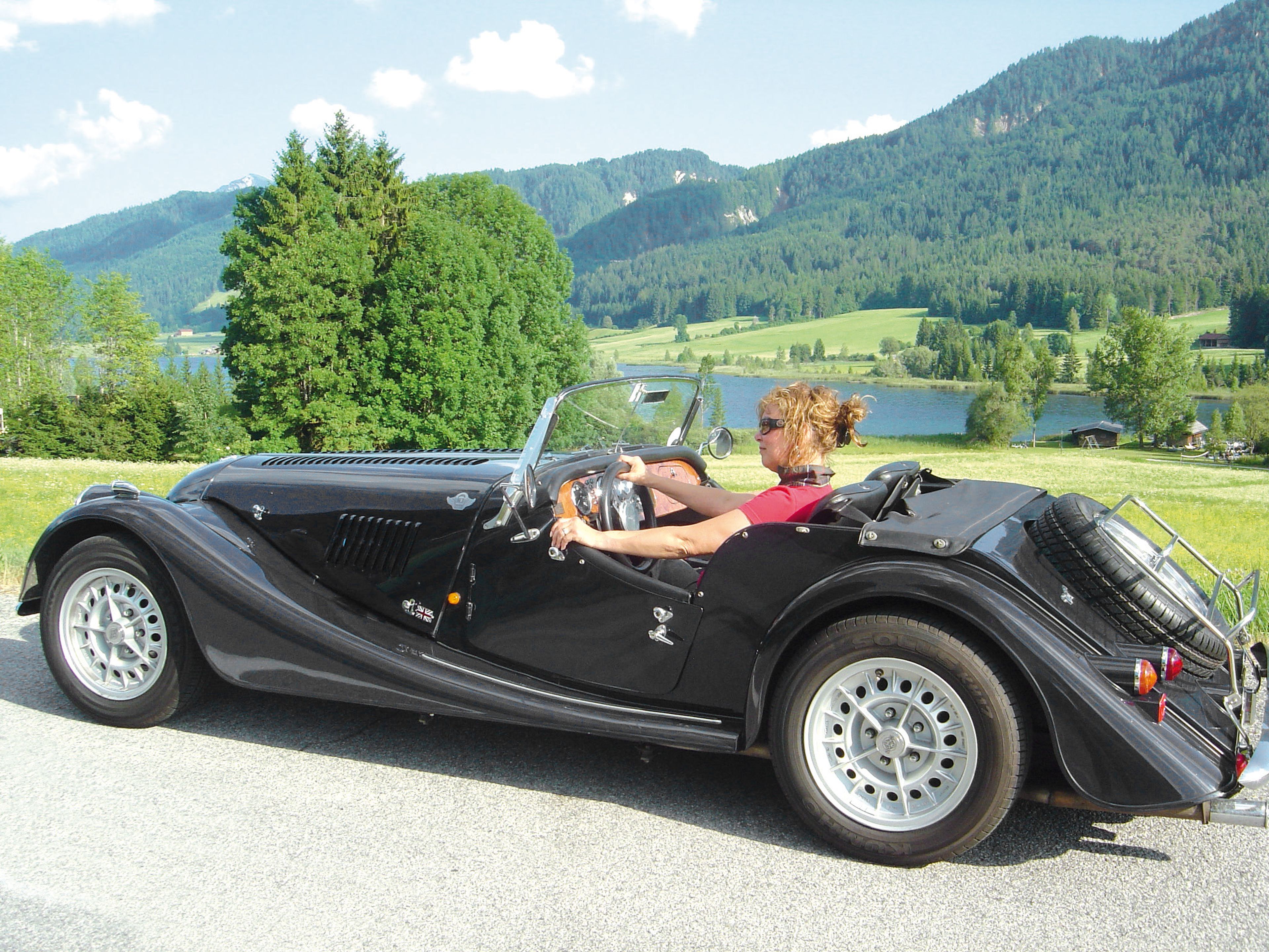 Frau sitzt in einem Oldtimer-Cabrio. Im Hintergrund der Weissensee und die Berge.