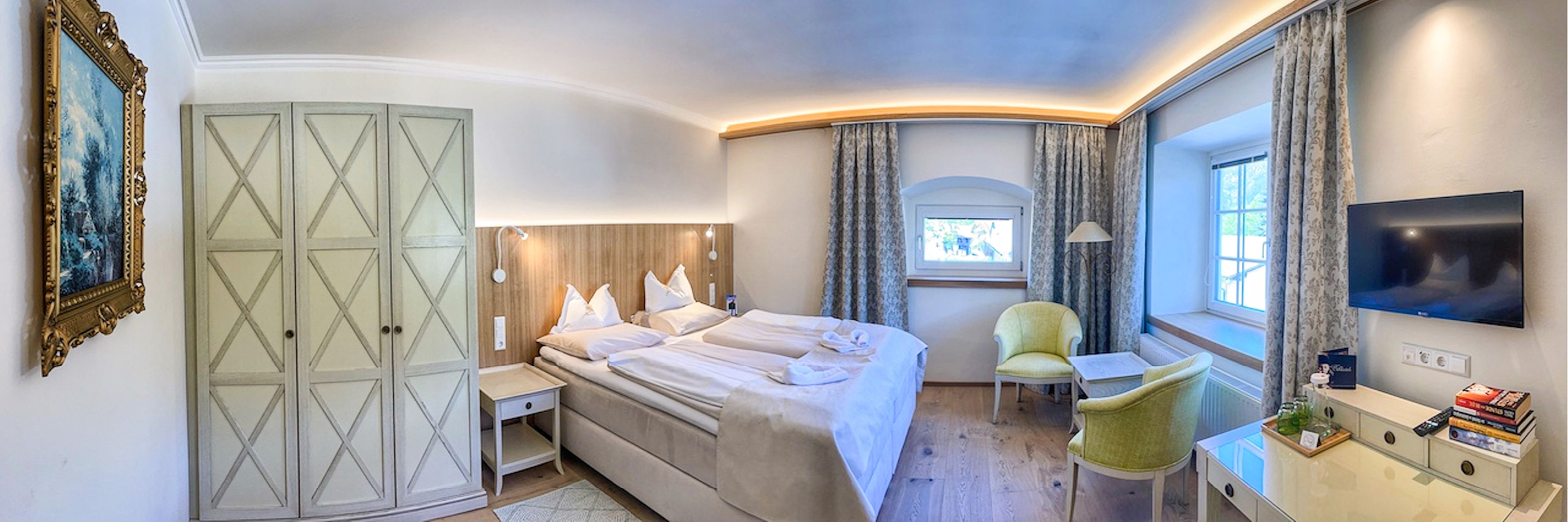 Cozy hotel room with a large bed, wooden floors, a TV, armchair, and window with curtains. Soft lighting and a framed painting on the wall.