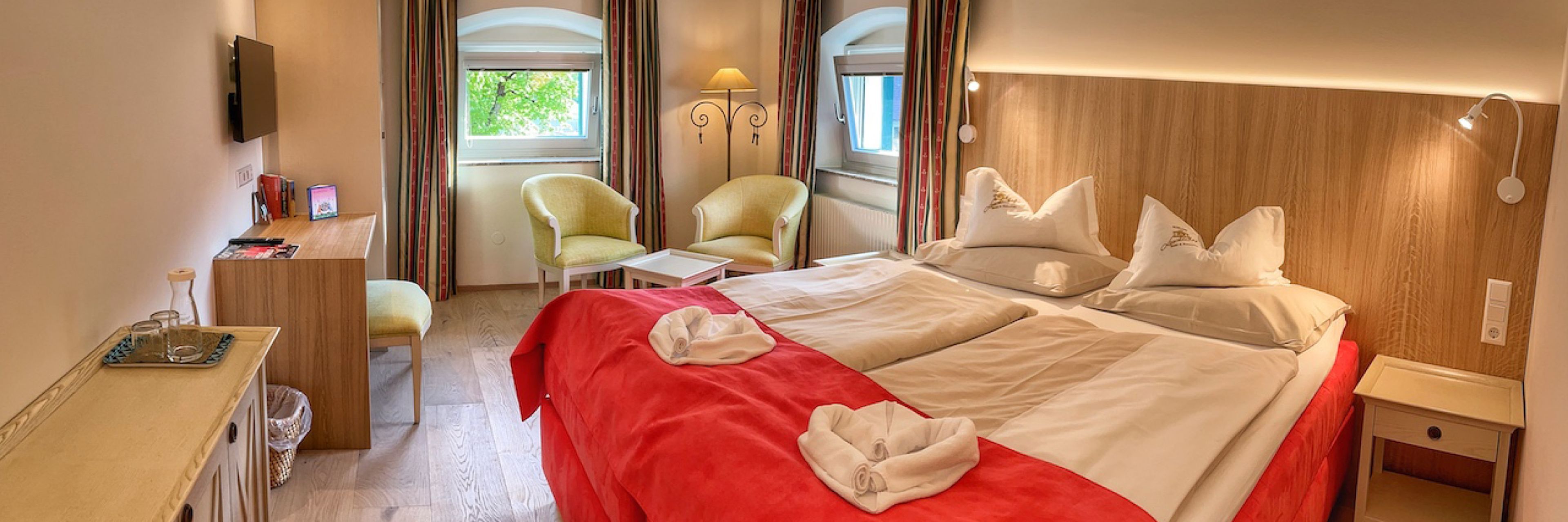 Cozy hotel room with a double bed, red blanket, two chairs, a desk, and a wall-mounted TV. Soft lighting and two windows with curtains.