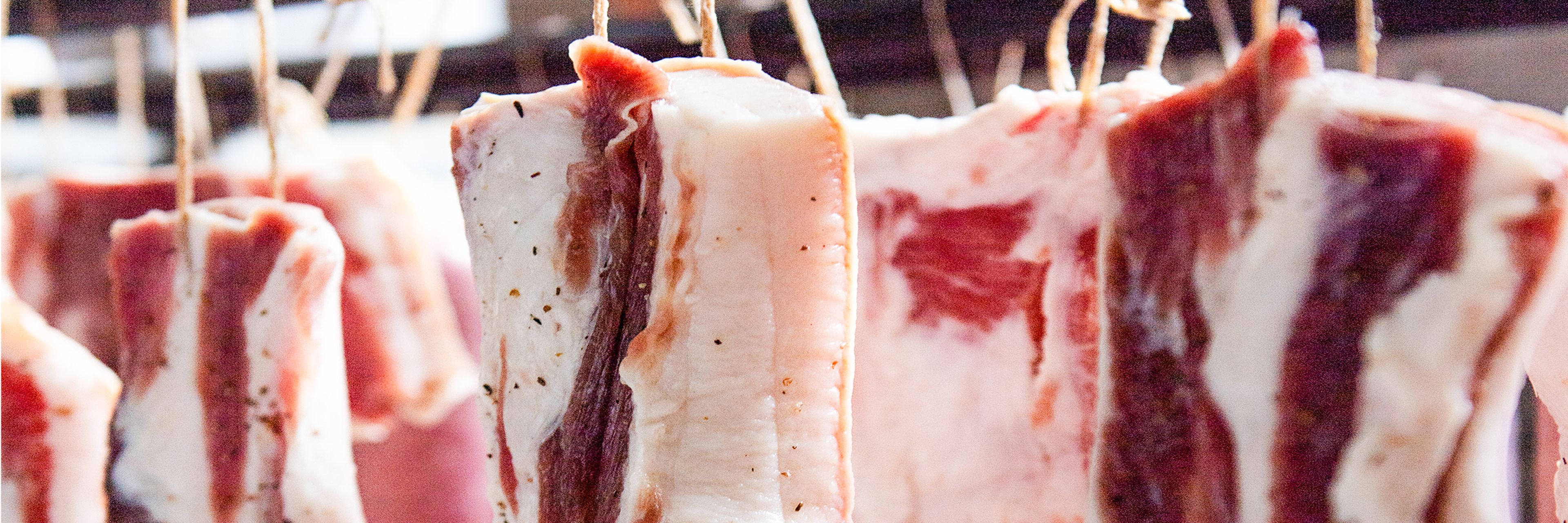 Nahaufnahme von vertikal an Schnüren hängenden Speckscheiben, die das marmorierte Fett und Fleisch in einem Reife- oder Trocknungsprozess zeigen.