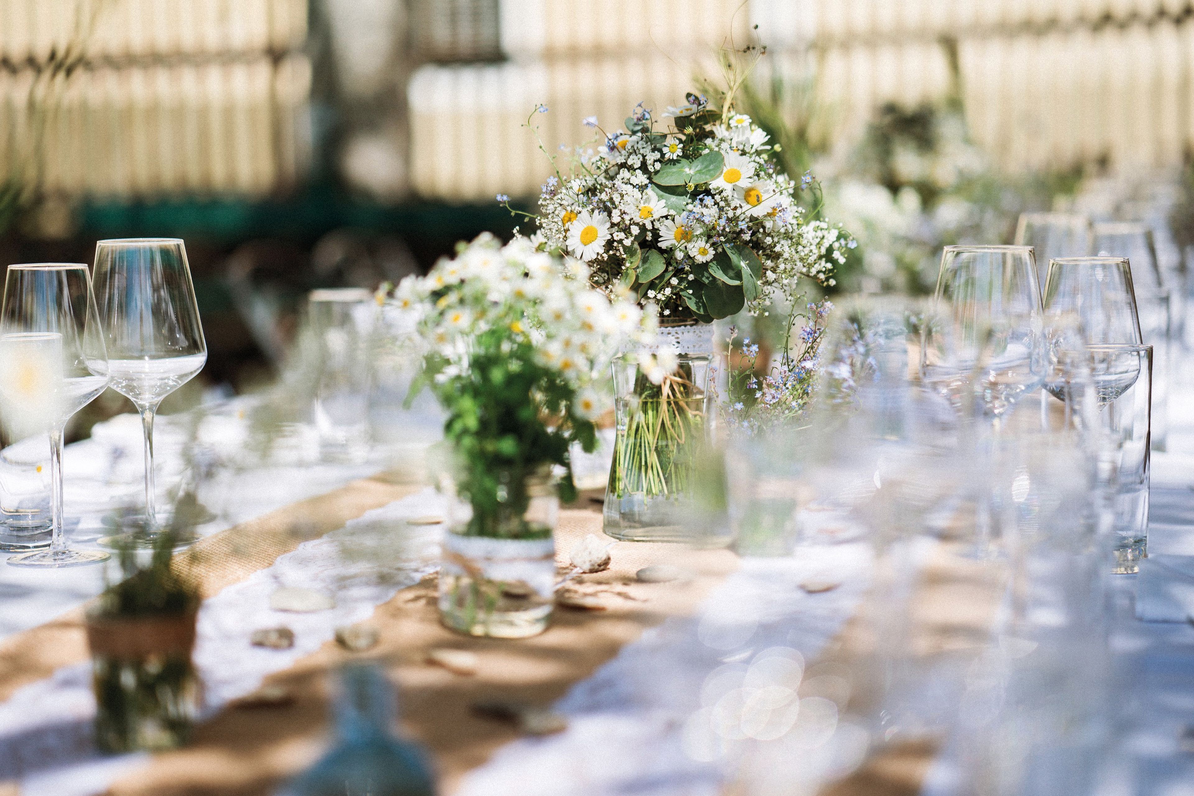 Ein für eine Veranstaltung im Freien gedeckter Tisch mit Weingläsern, einem Läufer aus Sackleinen und floralen Mittelstücken aus Gänseblümchen und Grünzeug.