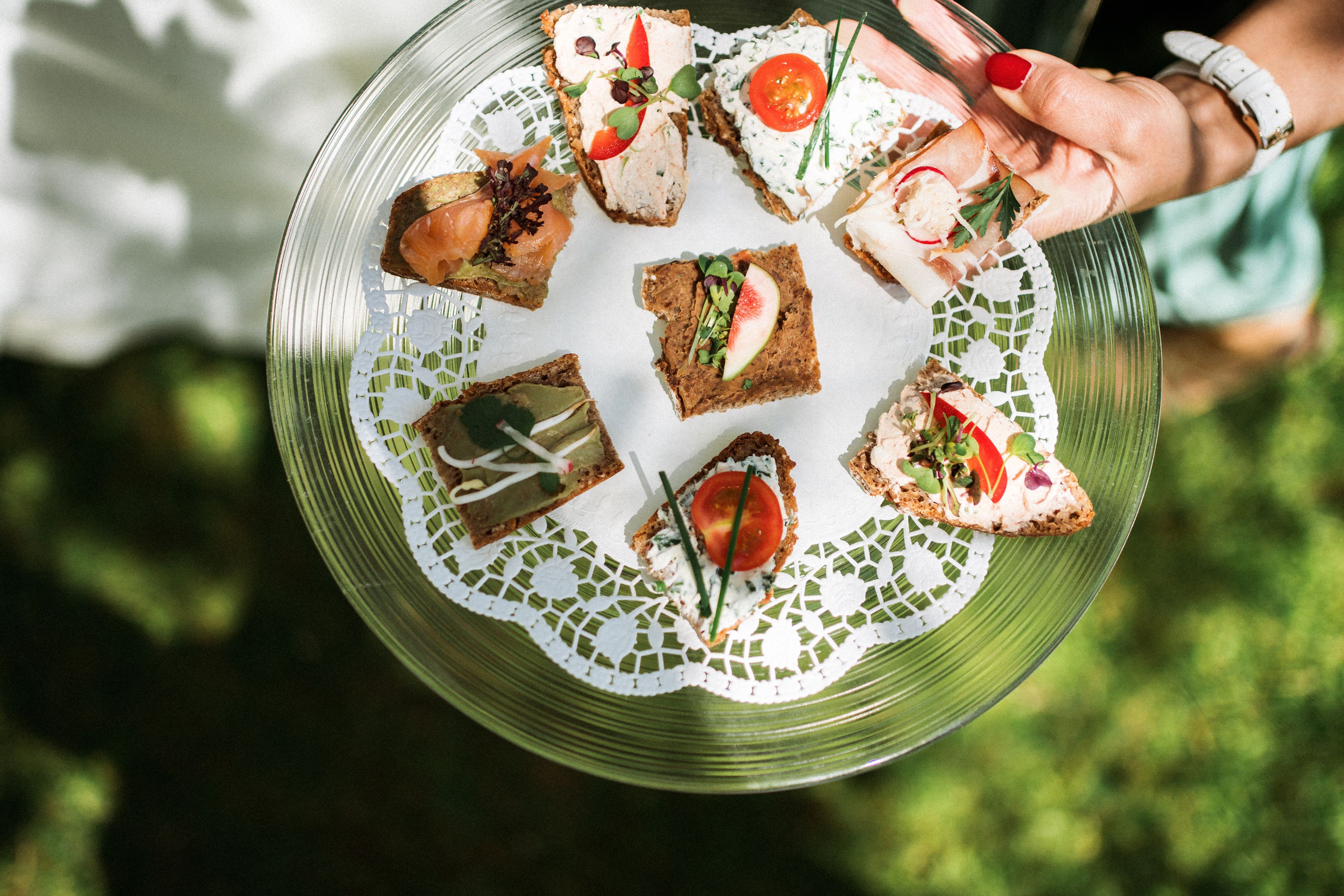 A hand holds a glass platter with assorted open-faced sandwiches, garnished with tomatoes, radishes, herbs, and a lace doily underneath.