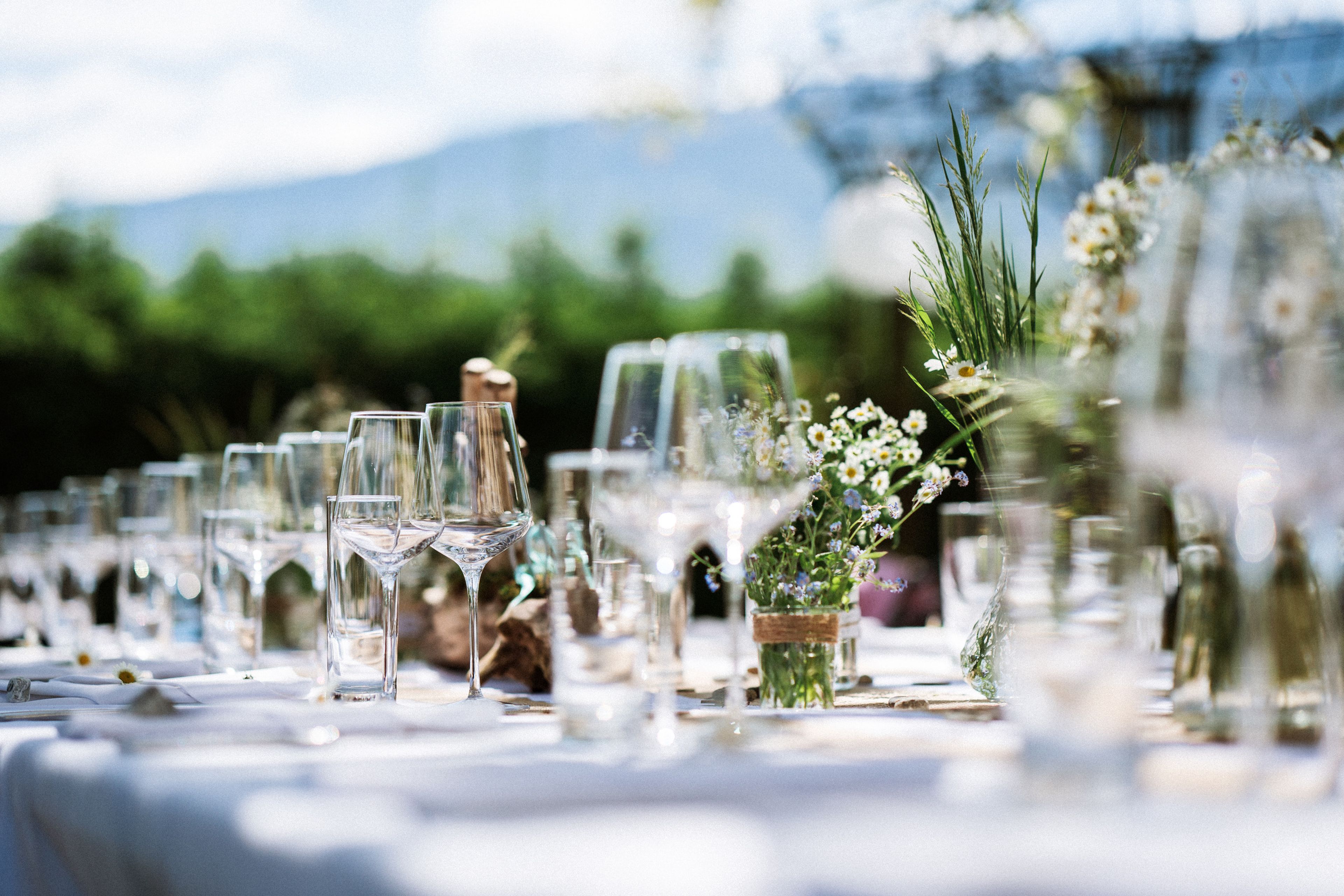 Elegant outdoor table setting with clear wine glasses, white tablecloth, and small floral centerpieces under a sunny sky.