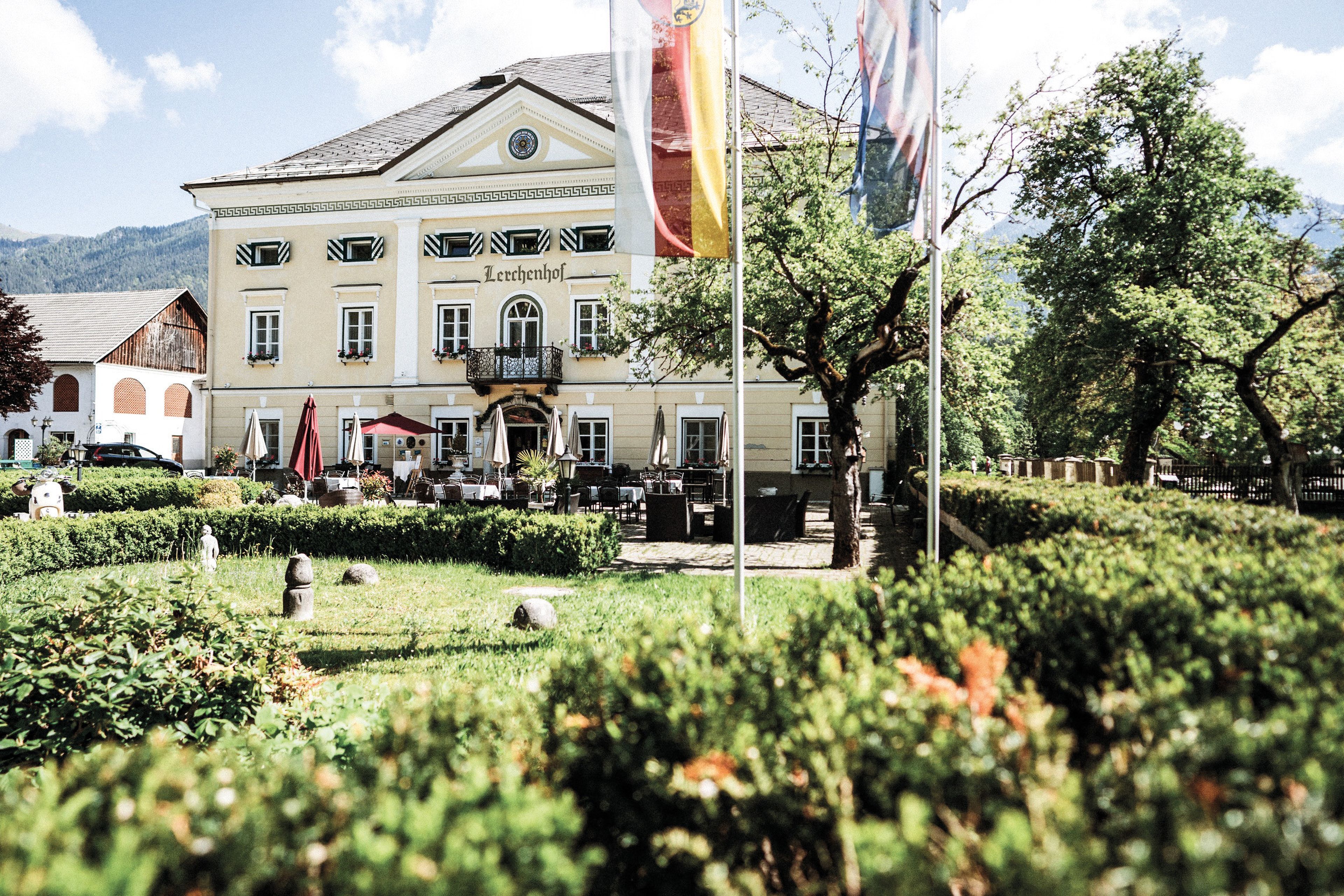 Das Schloss mit gelber Fassade und Balkon, umgeben von einem üppigen Garten mit Hecken und Fahnen, vor einer Bergkulisse.