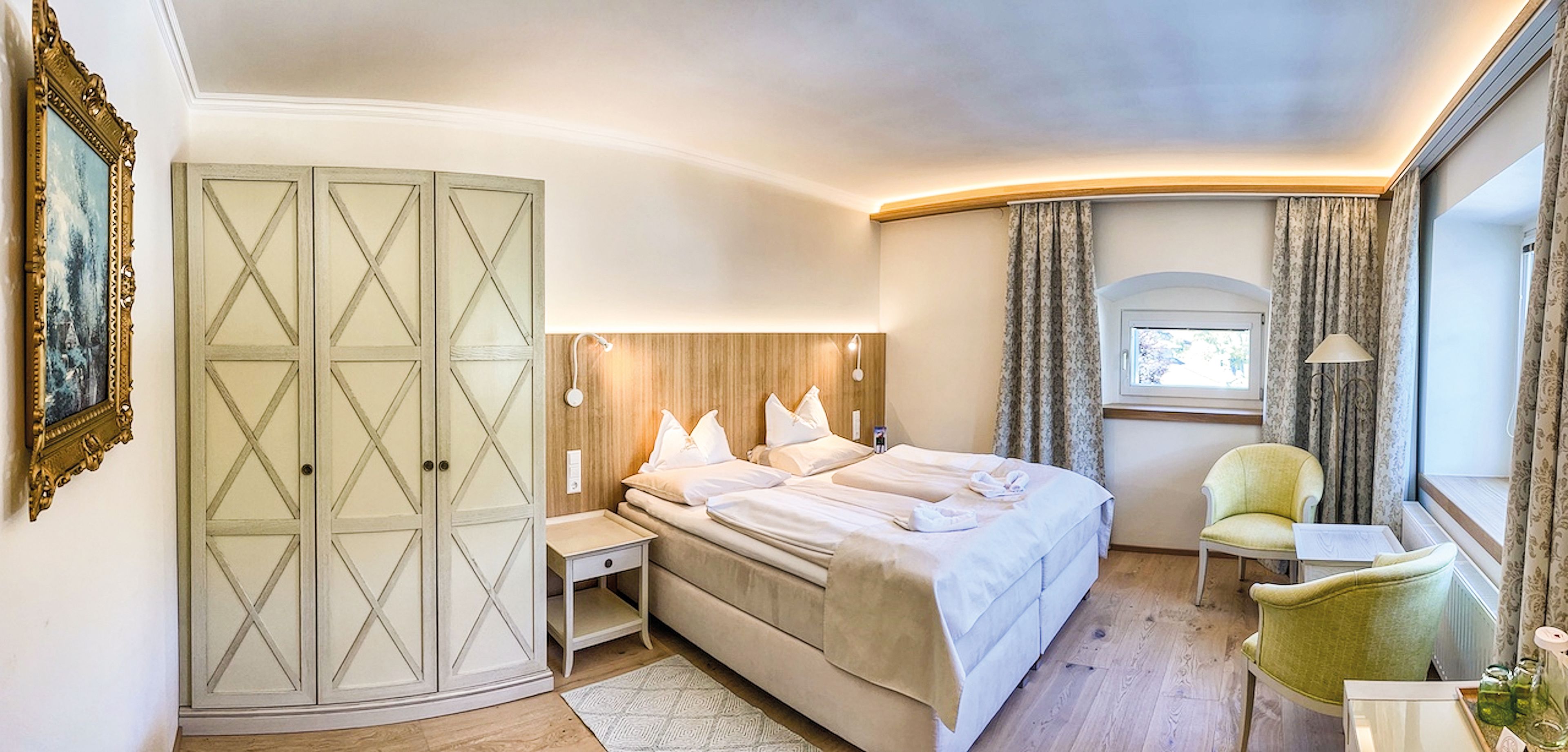 Elegantes Hotelzimmer mit einem großen Bett, gemütlichen Stühlen, einem Fenster mit Vorhängen, einem Kleiderschrank und einem Gemälde an der Wand. Die warme Beleuchtung wertet den Raum auf.
