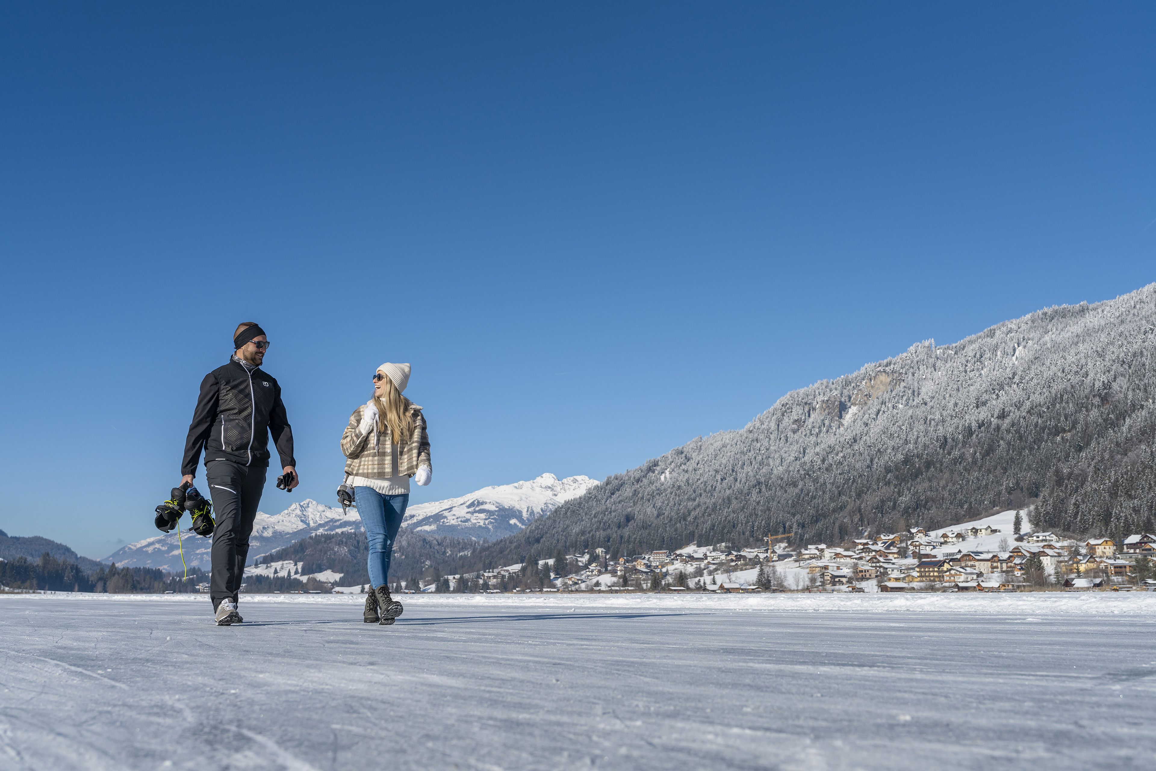 Ein Mann und eine Frau gehen mit Schlittschuhen auf einem zugefrorenen See spazieren, mit schneebedeckten Bergen und einem Dorf im Hintergrund unter einem klaren blauen Himmel.