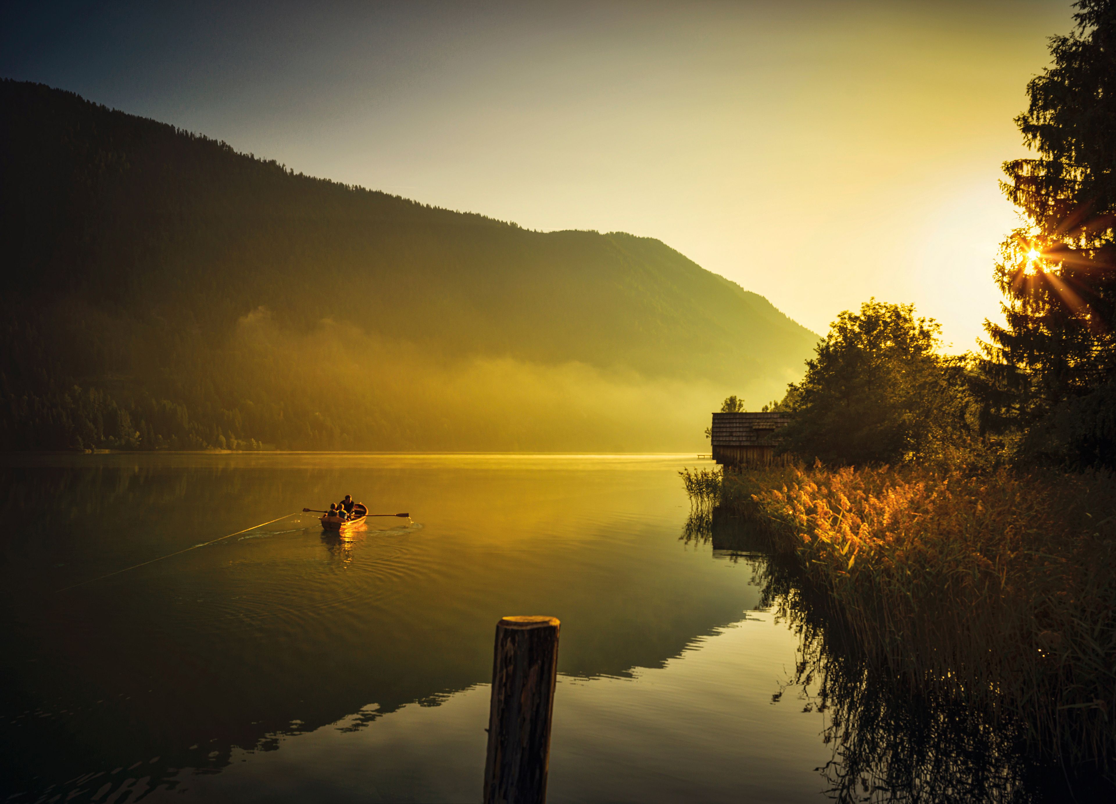 Ein kleines Boot auf einem ruhigen See bei Sonnenuntergang, umgeben von Bergen und Bäumen, mit goldenem Licht, das sich auf dem Wasser spiegelt.