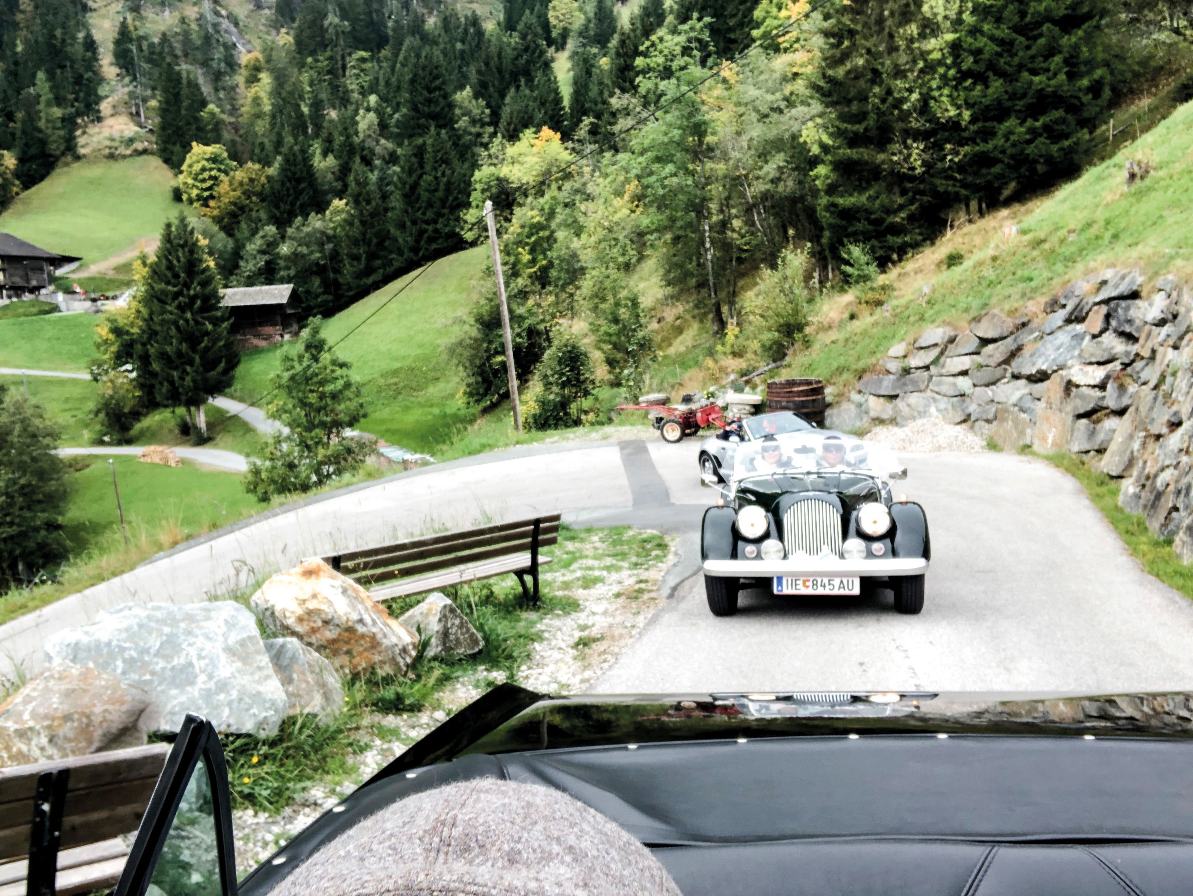 Oldtimer fahren auf einer schmalen Bergstraße, umgeben von üppigem Grün und Bäumen, mit einer Steinmauer auf der rechten Seite.