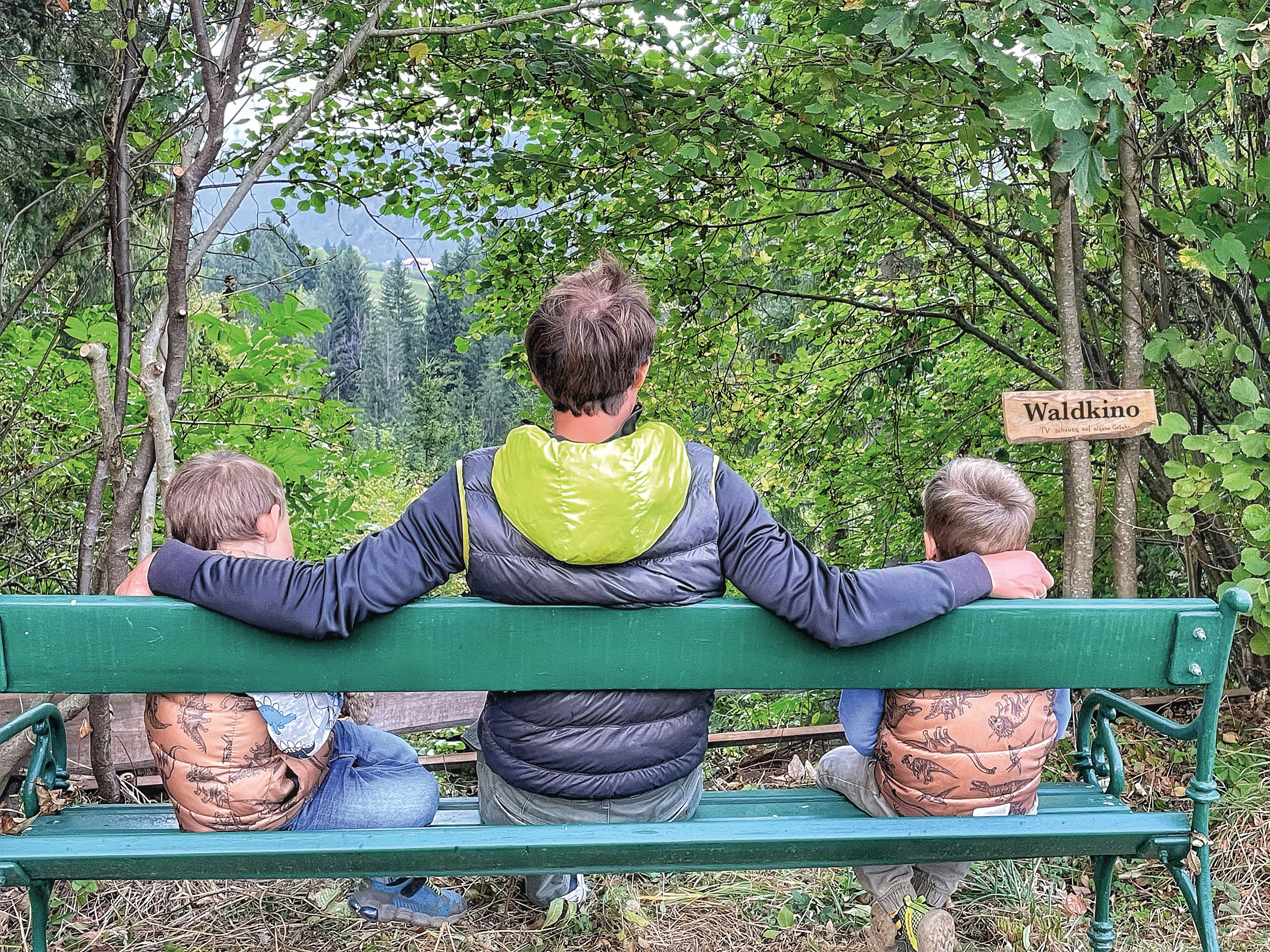 Ein Mann sitzt auf einer grünen Bank im Wald und hat seine Arme um zwei Kinder gelegt. Im Hintergrund ist dichter Wald zu sehen. Rechts hängt ein Holzschild mit der Aufschrift 'Waldkino'