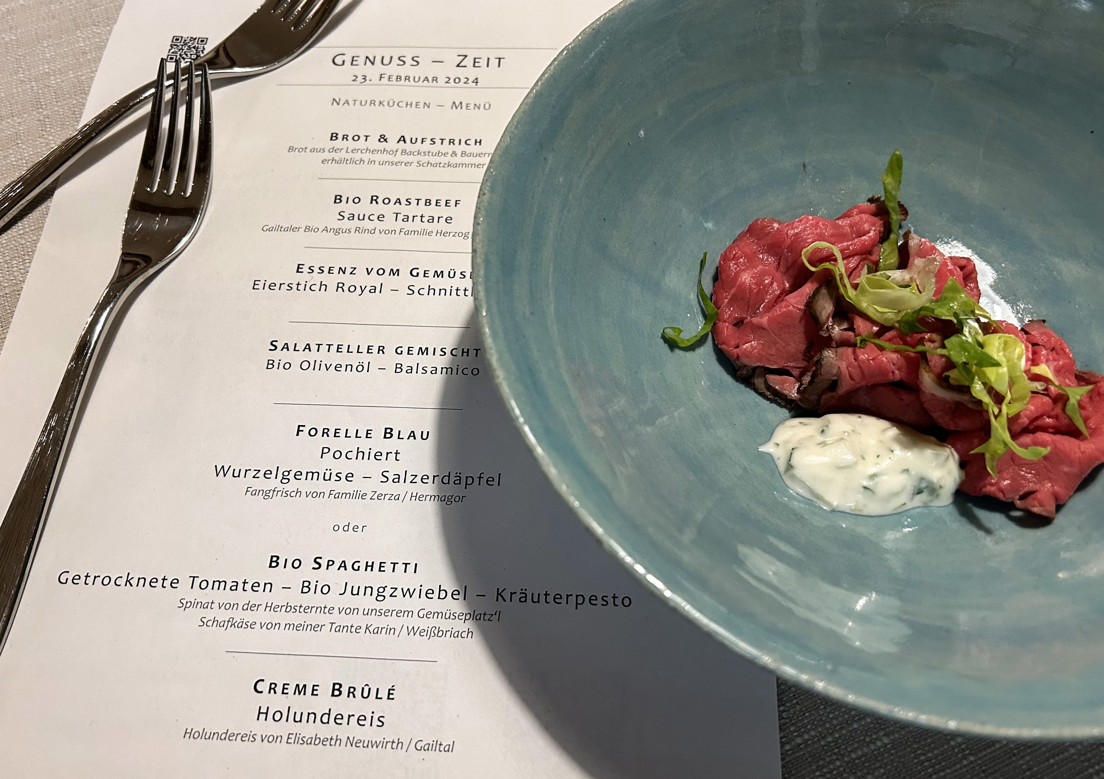 A menu titled "Genuss – Zeit" with a dish of thinly sliced roast beef, greens, and sauce on a blue plate. Silverware is visible.