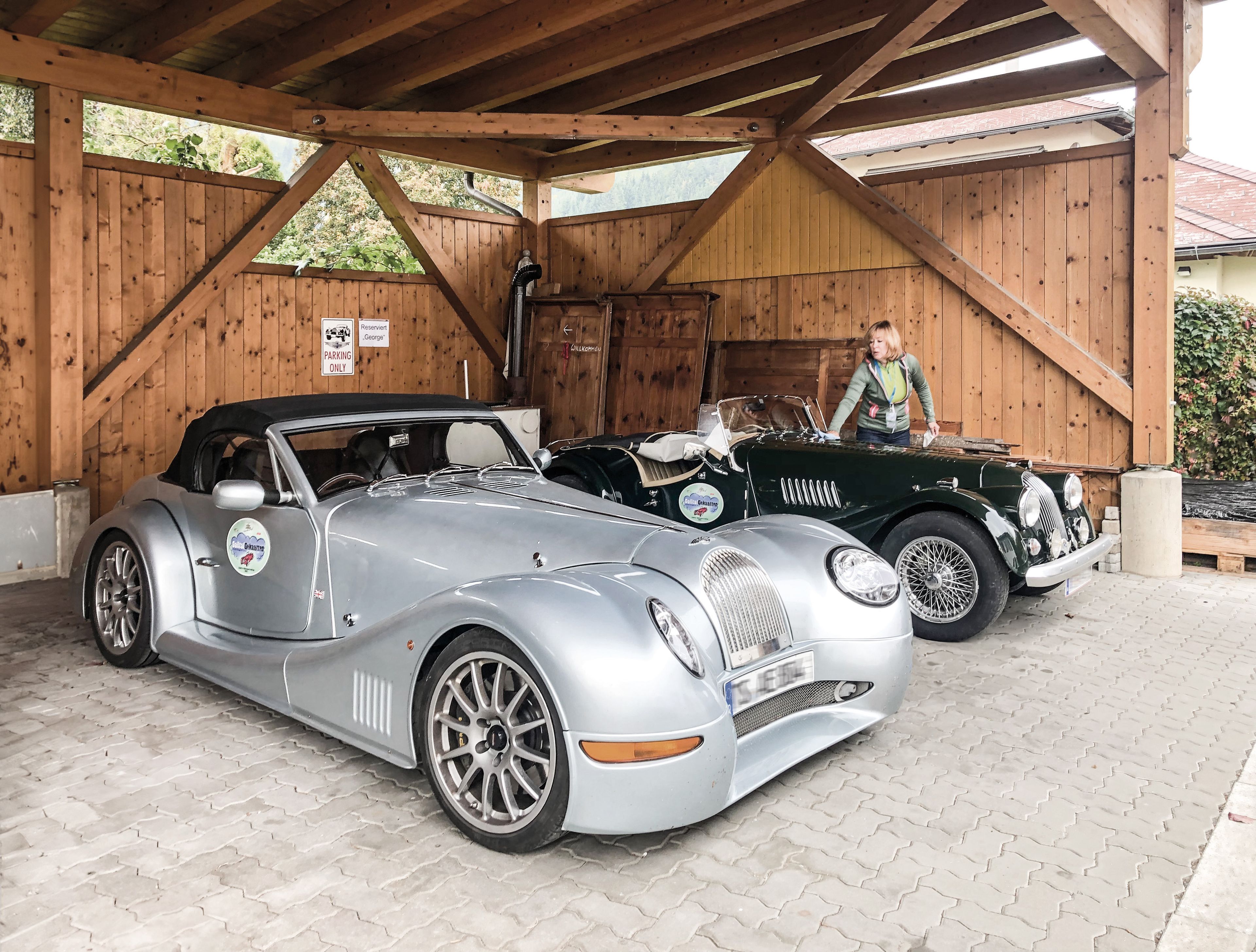 Two vintage-style sports cars, one silver and one green, parked under a wooden shelter. A person stands beside the green car.