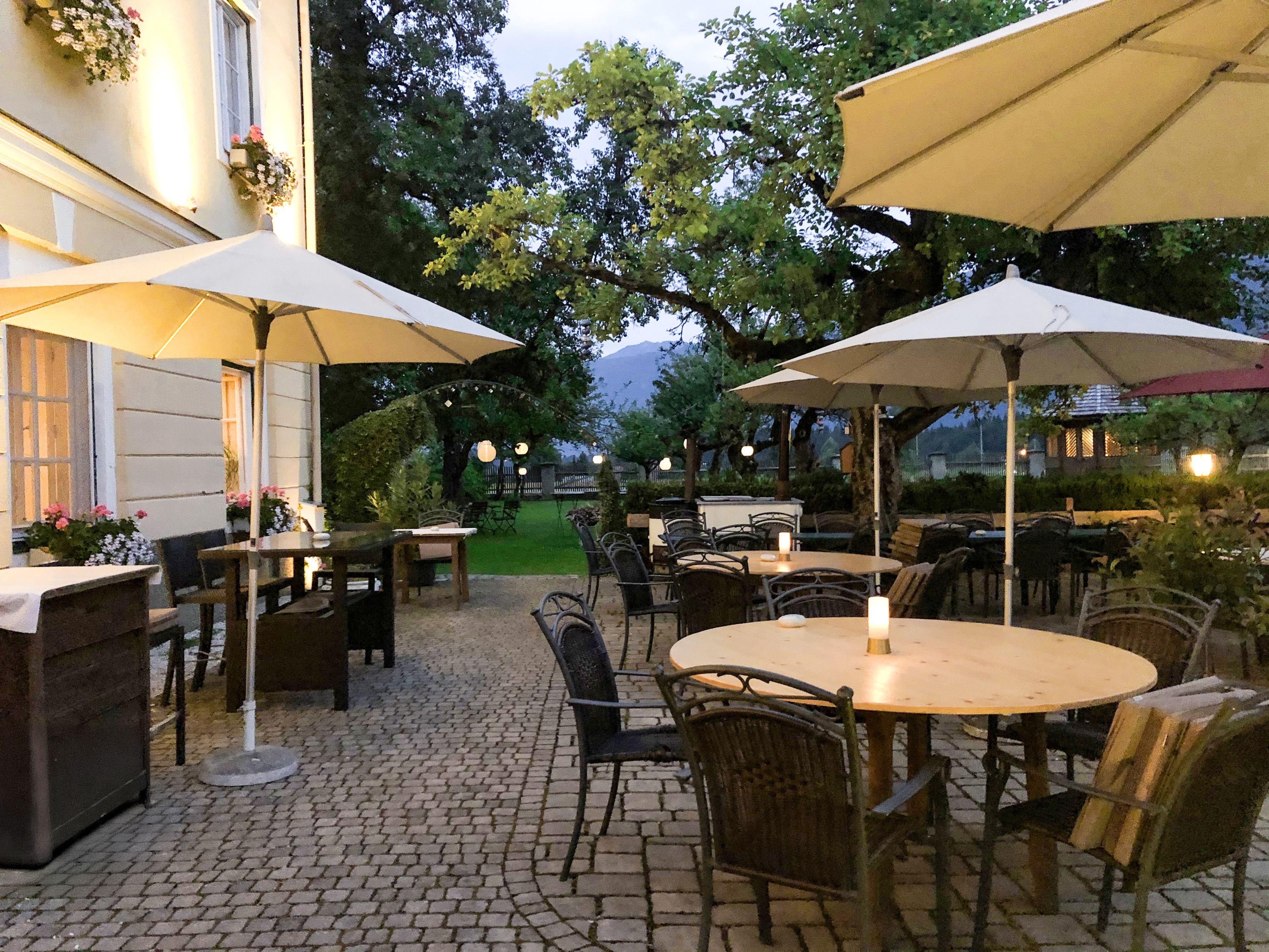 Restaurantterrasse im Freien mit runden Tischen, Korbstühlen und großen Sonnenschirmen. Warmes Licht und viel Grün schaffen eine gemütliche, einladende Atmosphäre.