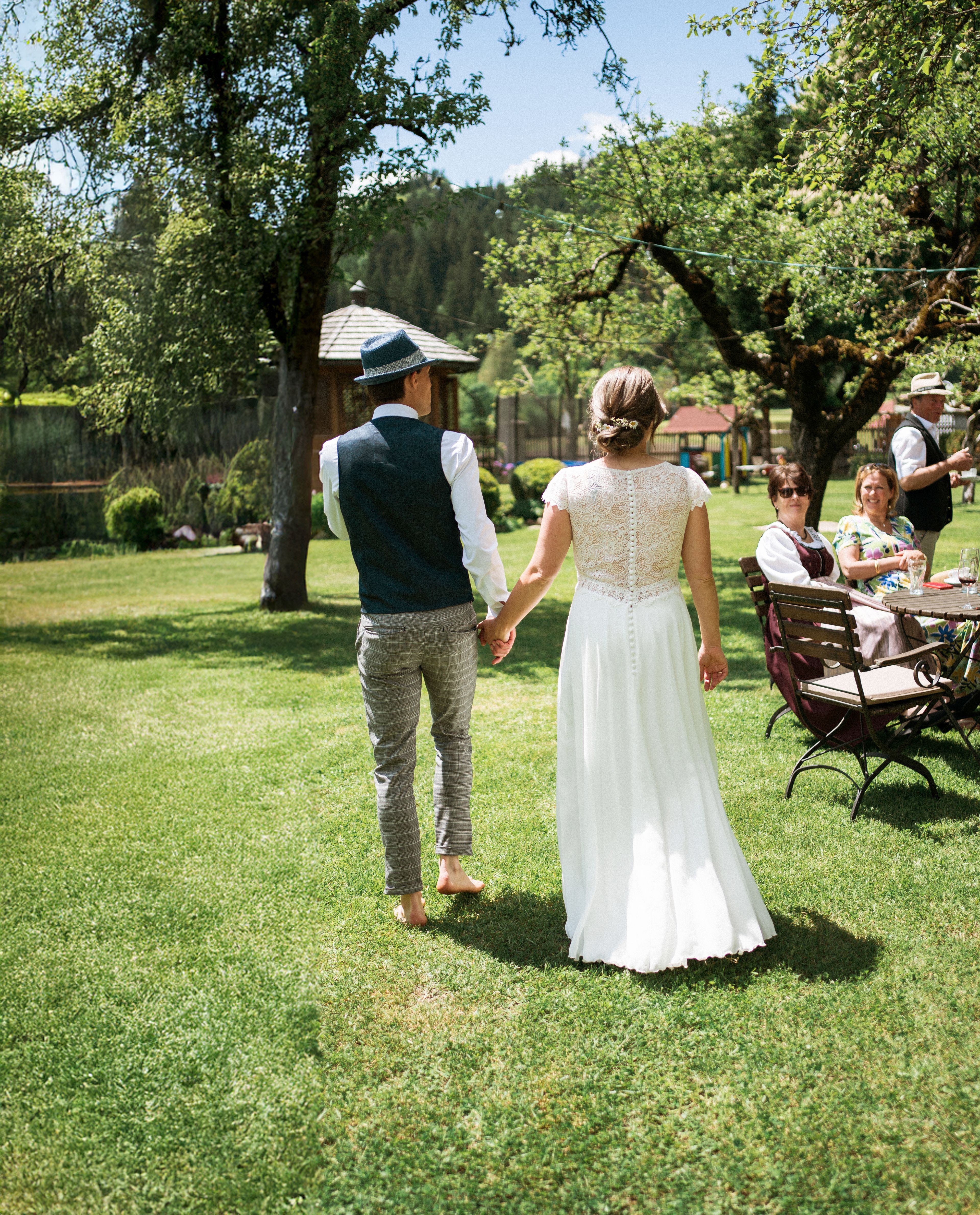 Hochzeitspaar läuft durch den Garten des Schloss Lerchenhofs, barfuß in der sommerlichen Wiese, Gäste lächeln ihnen sitzend zu