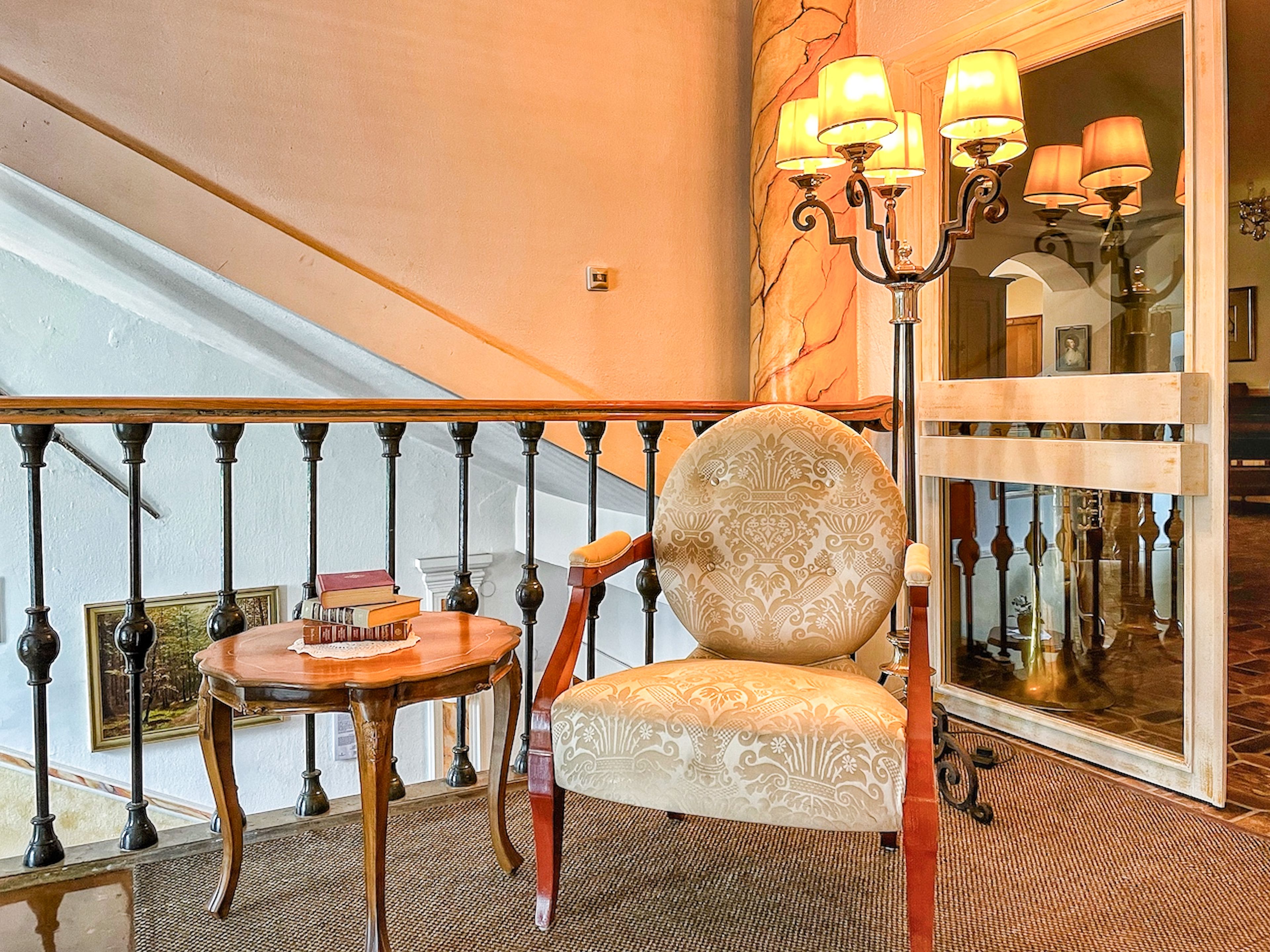 Eleganter Sitzbereich mit gemustertem Sessel, Holztisch mit Büchern und hoher Lampe vor einer Spiegelwand und einem Treppengeländer.