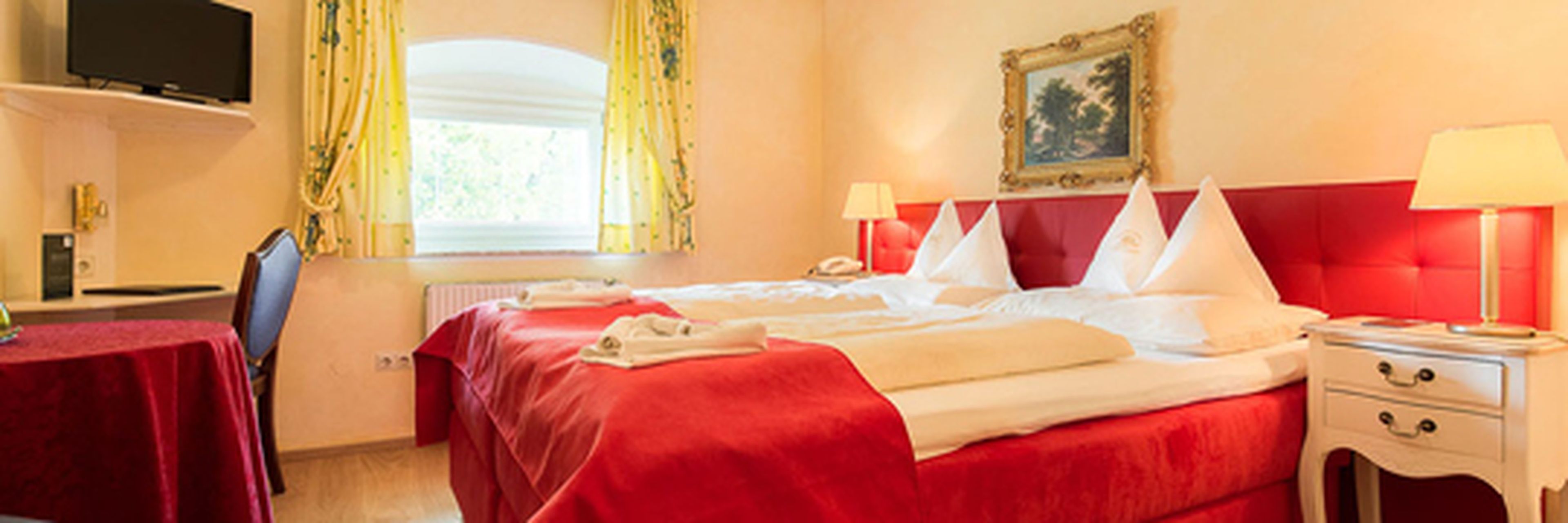 Cozy hotel room with two beds, red headboards, matching bedspreads, two bedside lamps, a wall painting, a TV, and a window with yellow curtains.
