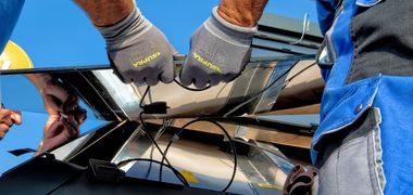 Zwei Menschen heben mit Arbeitshandschuhen eine silberne, verkabelte Platte in eine Fassung.