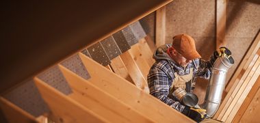 Ein Handwerker sitzt in einer Holzkonstruktion und hält ein großes gewundenes Rohr aus Metall.