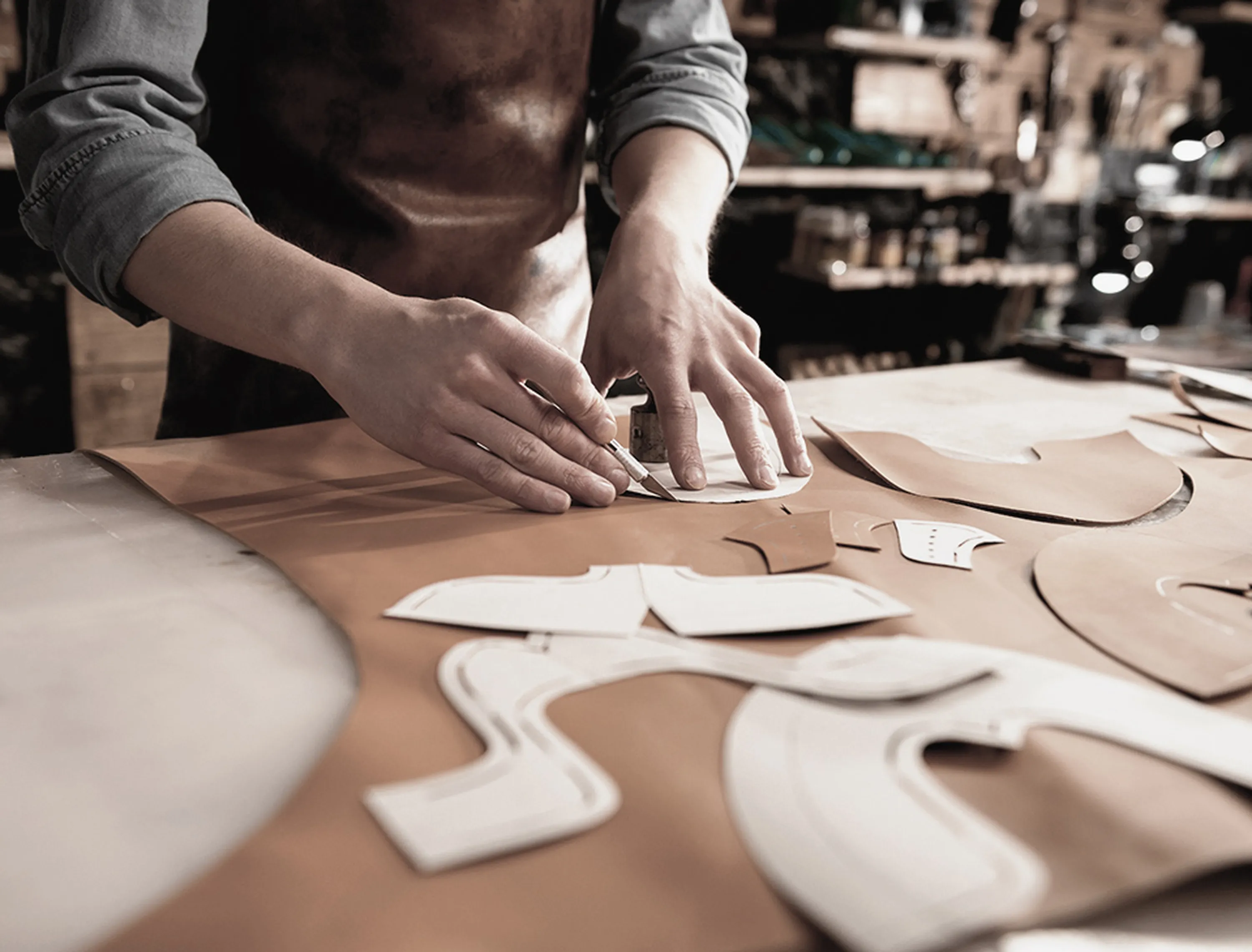 Person cutting leather pieces on a workbench, surrounded by various pattern templates in a workshop setting.