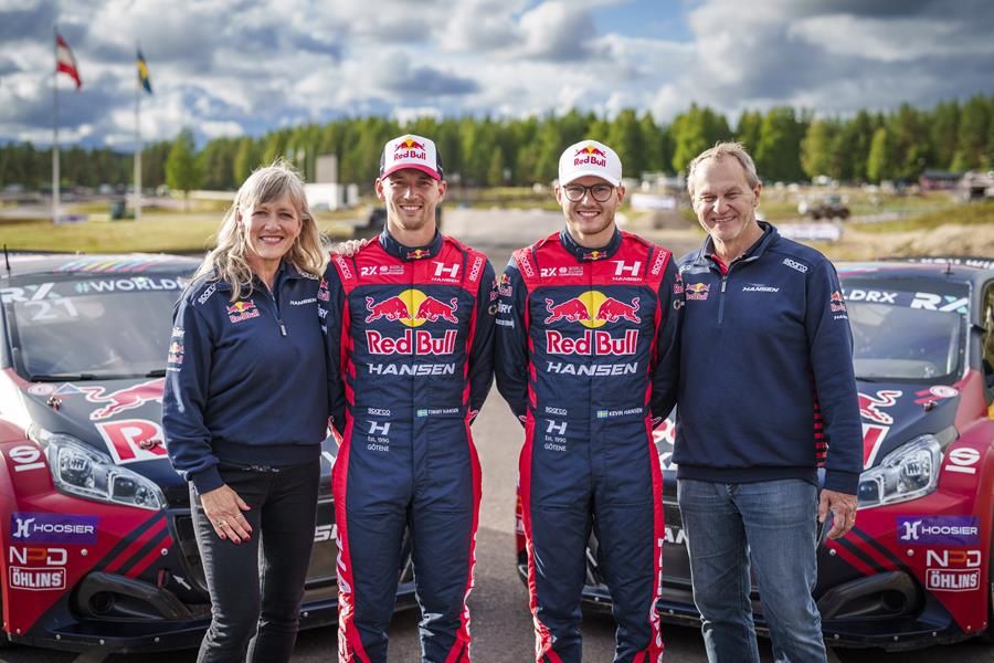 Four people stand in front of two rallycross racing cars smiling