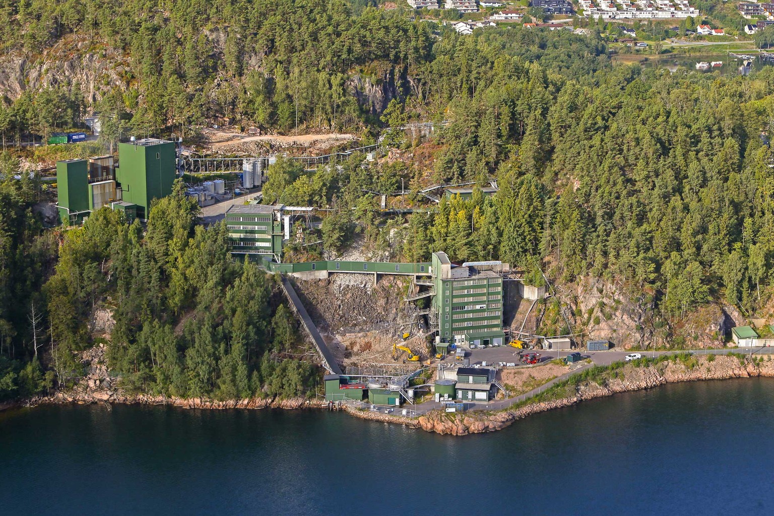 Aerial view of an industrial facility nestled in a forested hillside by a calm body of water, with several green buildings and construction equipment.
