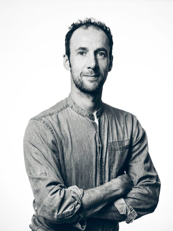 Black and white portrait of a man with short curly hair, wearing a button-up shirt, standing with arms crossed against a plain background.