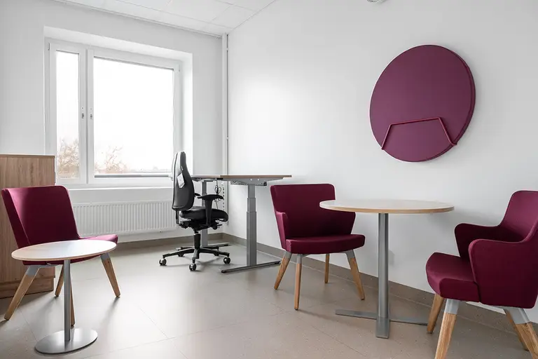 Modern office interior with purple chairs, round tables, and an adjustable black office chair by a window. A circular wall panel decorates the wall.