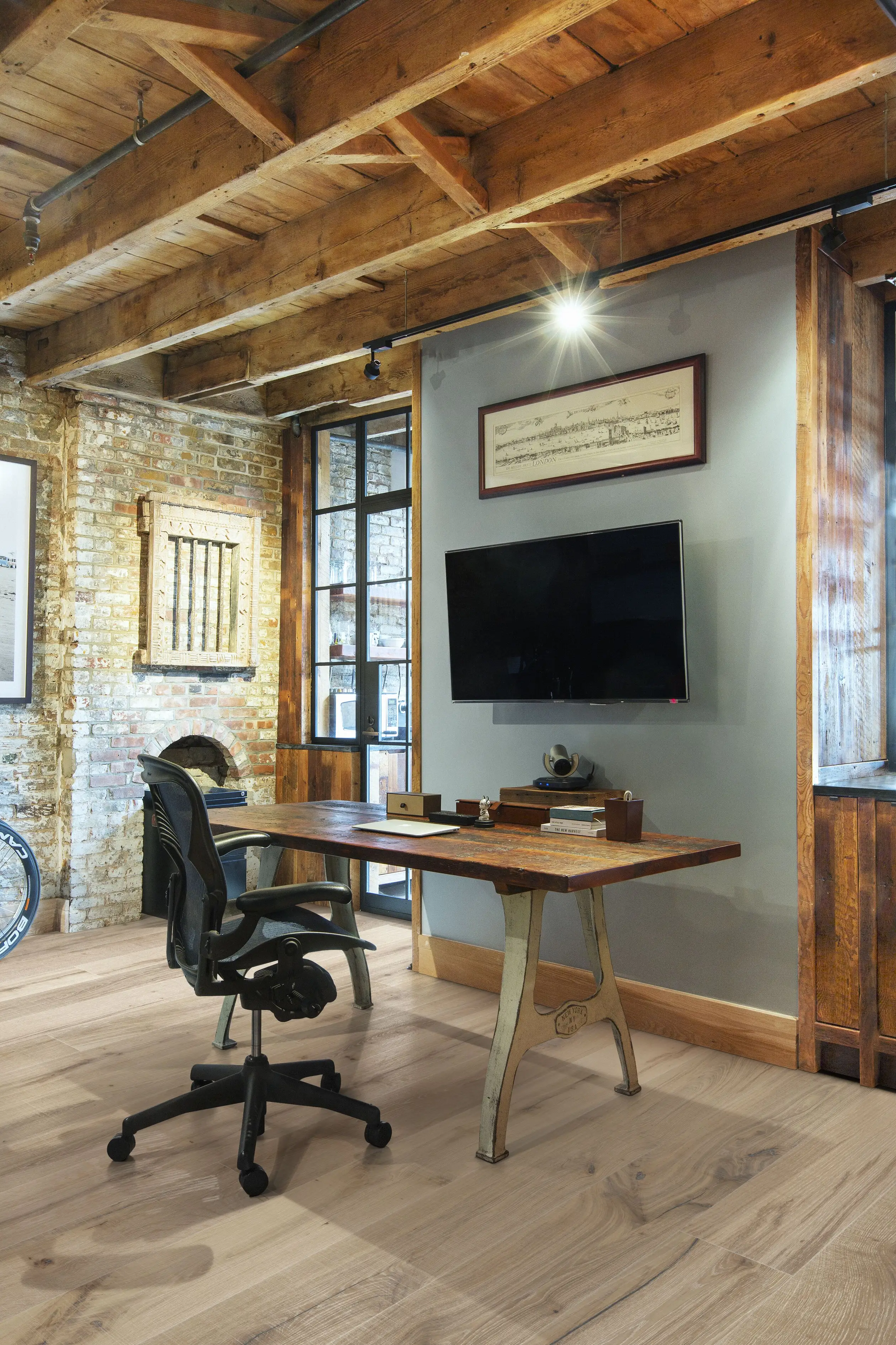 Rustic home office with a wooden desk, black chair, exposed brick walls, wooden ceiling beams, and a wall-mounted TV.